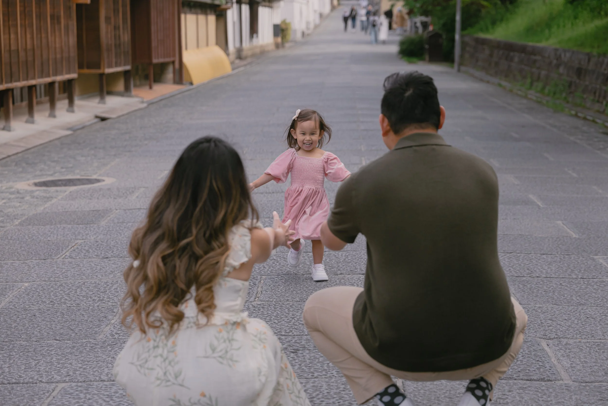 A young girl in a pink dress runs towards a seated man and woman on an outdoor city street, with other pedestrians in the background.