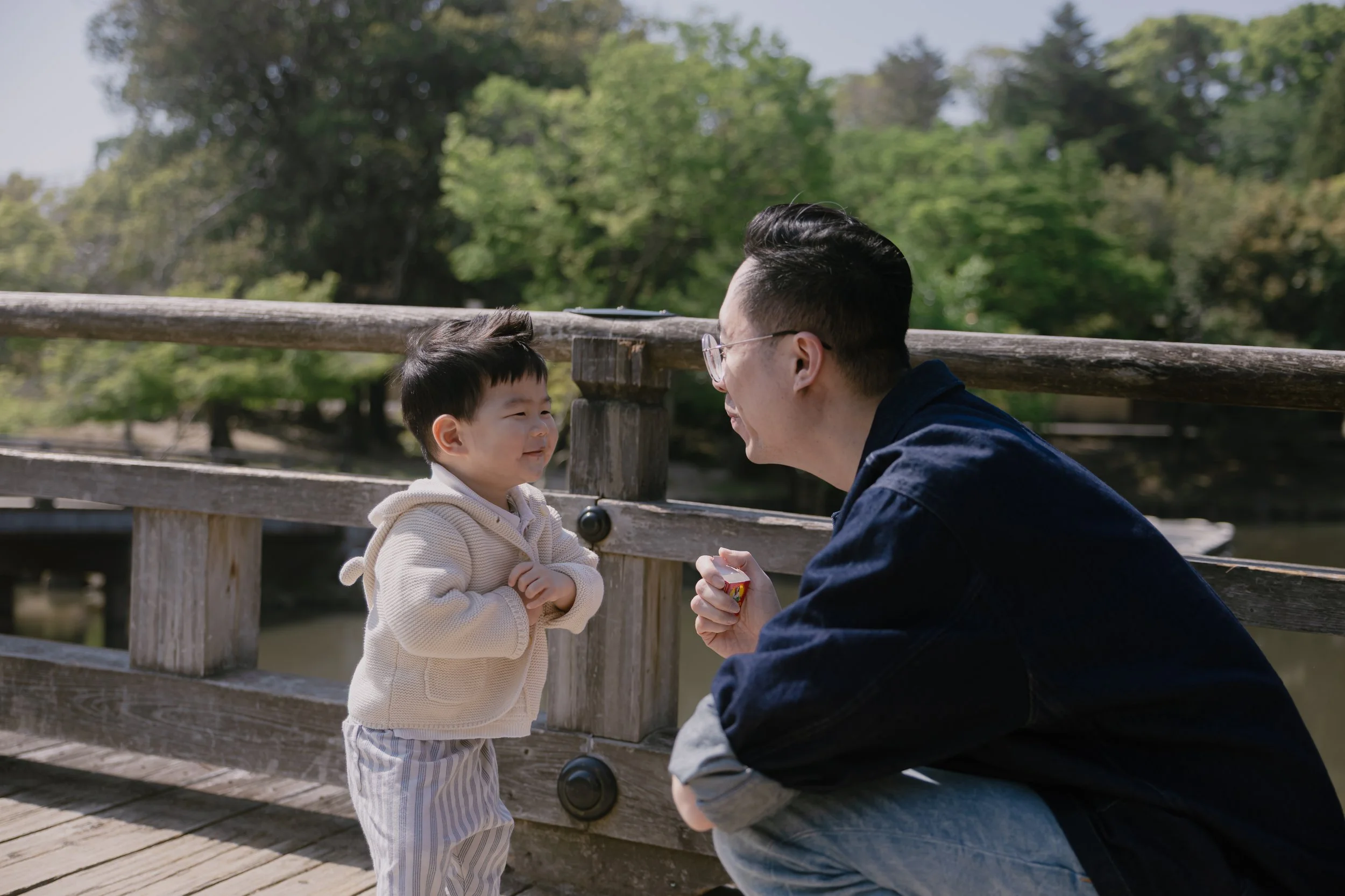 A man and a young boy are outdoors on a wooden bridge surrounded by green trees. The man is squatting, facing the boy, and holding something in his hands. The boy is standing, facing the man, and smiling.