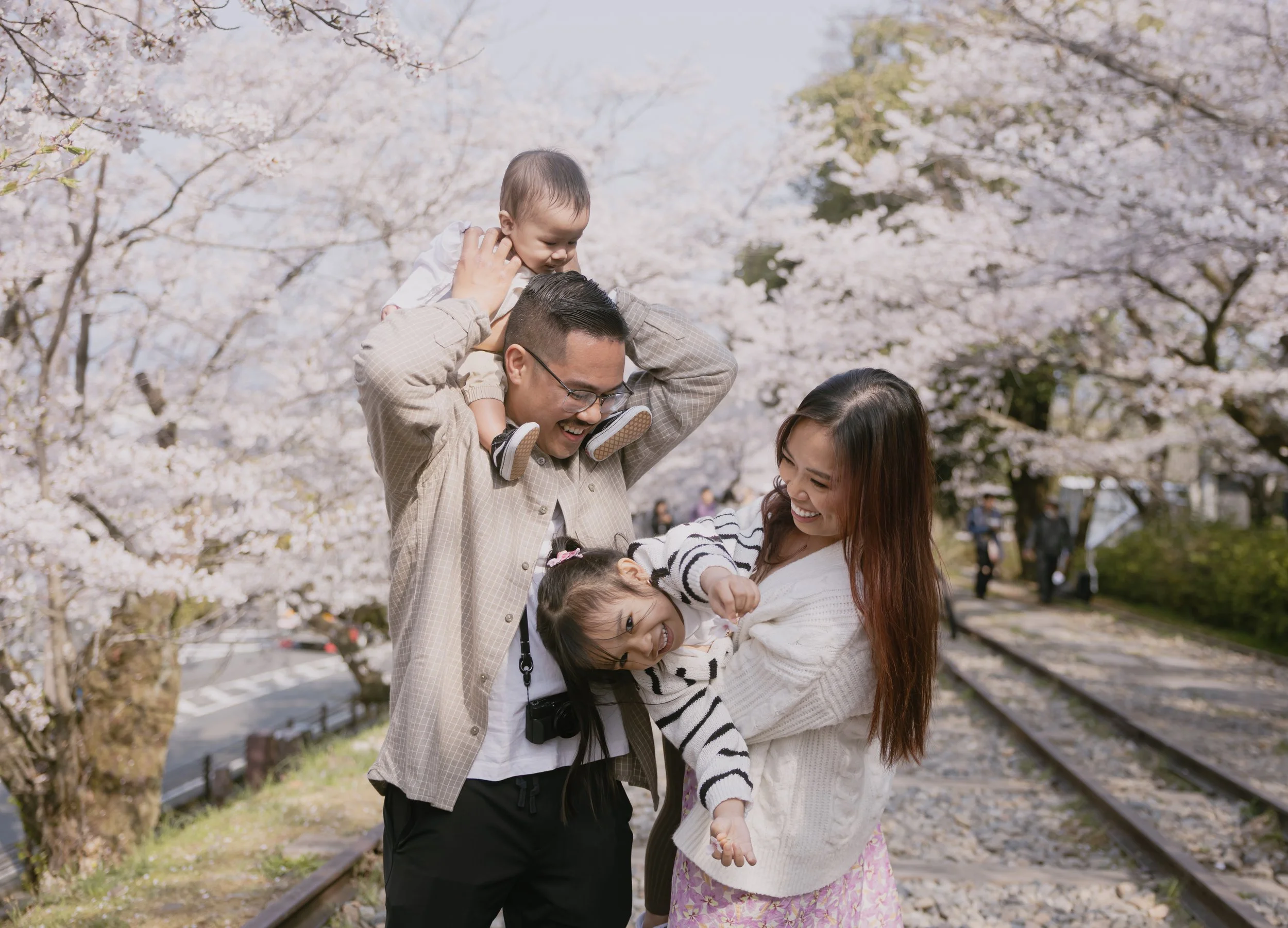 A family of four, including a man, woman, and two young girls, enjoying a walk under cherry blossom trees during springtime.