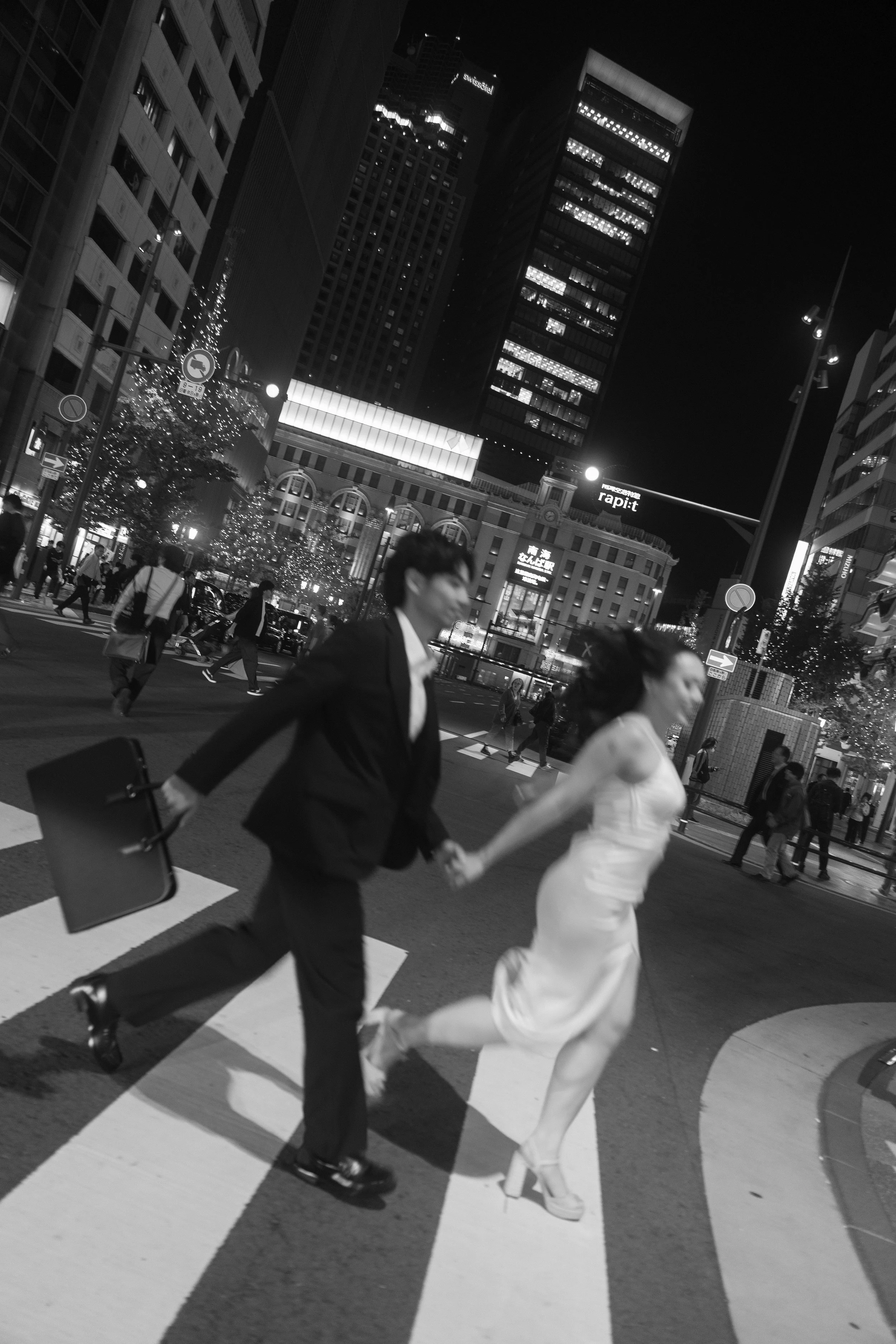 People crossing a city street at night, with tall buildings and illuminated signs in the background.