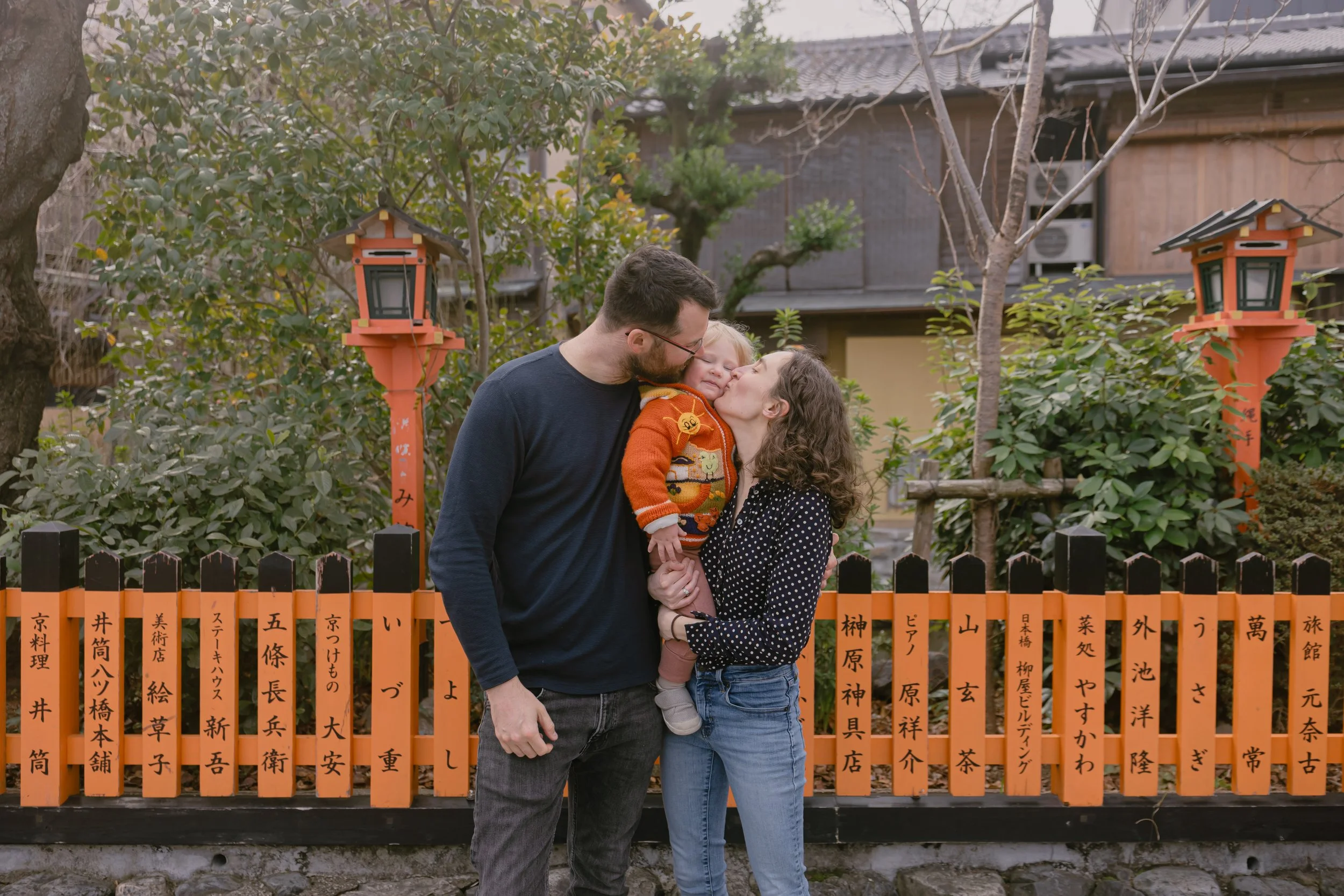 A family of three, a man, woman, and young girl, sharing a kiss outdoors in front of a traditional Japanese orange fence with black Japanese characters, surrounded by trees and greenery, in a garden with a building in the background.