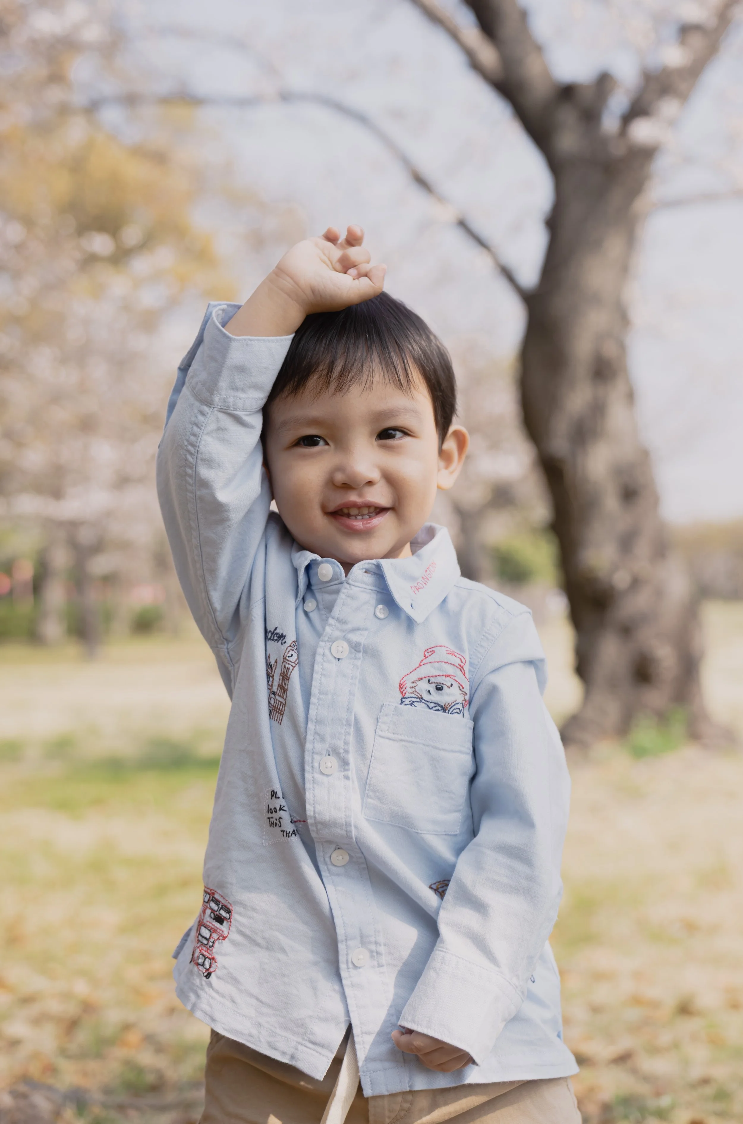 Young boy outdoors under a tree, smiling, raising his hand near his head on a sunny day.