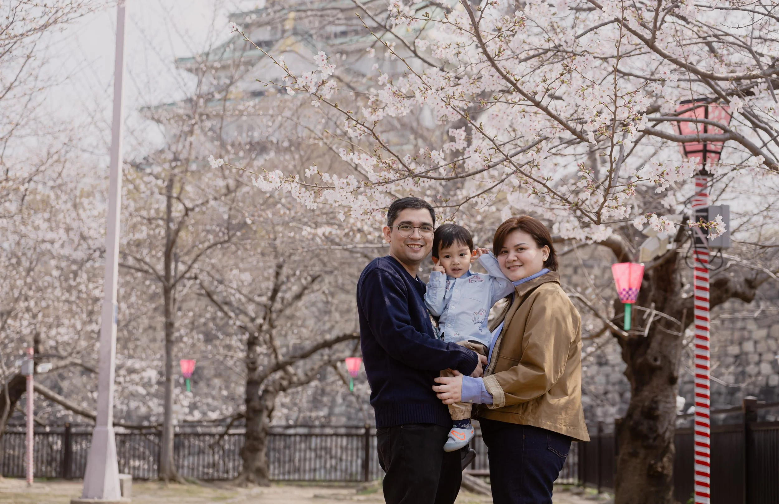 A family of three, consisting of a man, a woman, and a young child, stands together outdoors under blooming cherry blossom trees during spring time, with traditional paper lanterns hanging in the background.