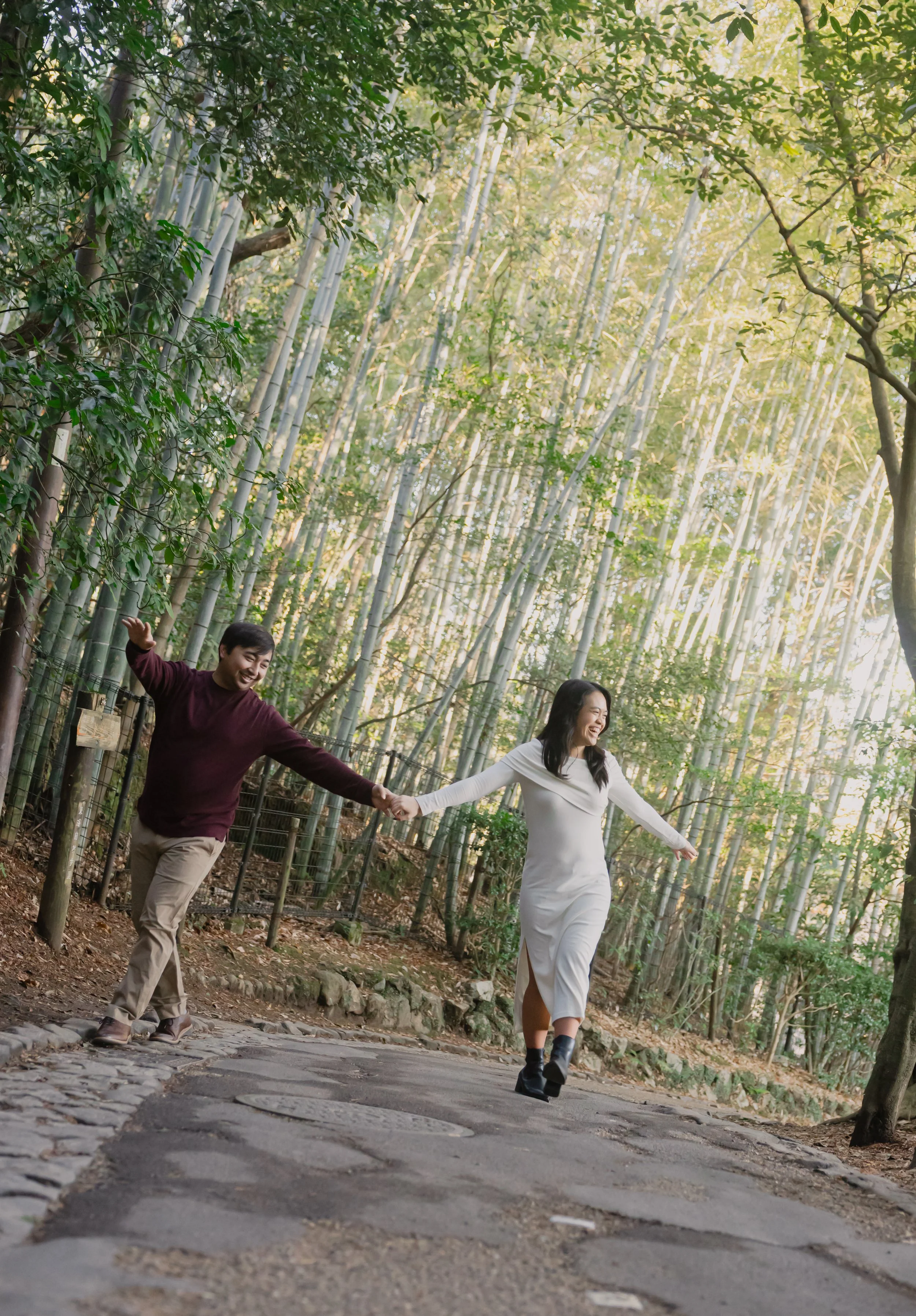 Two people walking along a forested path holding hands, smiling, with bamboo trees and sunlight in the background.