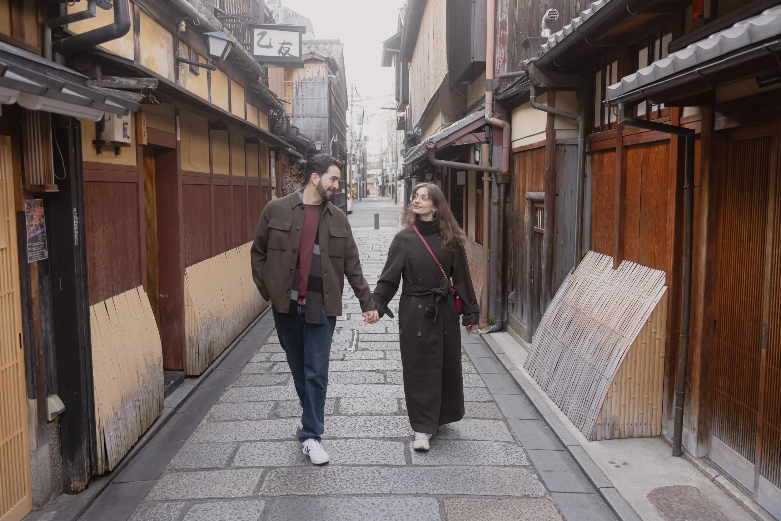 A man and woman holding hands walking down a narrow street with traditional Japanese wooden buildings on both sides.