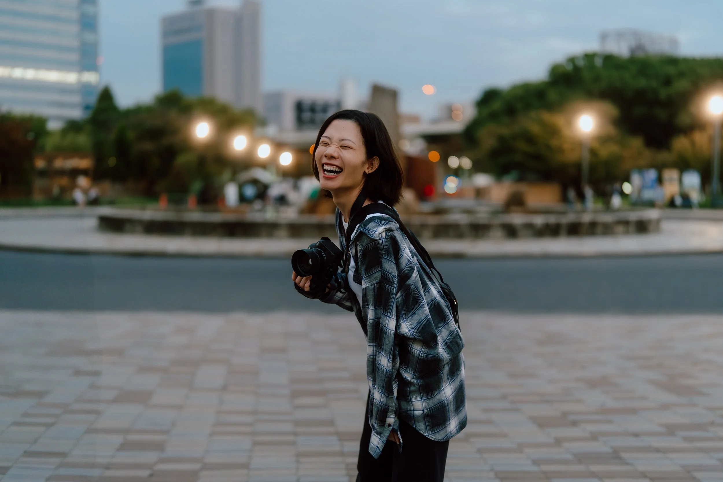 A woman with short black hair, wearing a plaid shirt, is smiling and laughing while holding a camera in her hand. She is standing outdoors in an urban park with buildings and trees in the background, illuminated by ambient streetlights in the evening.