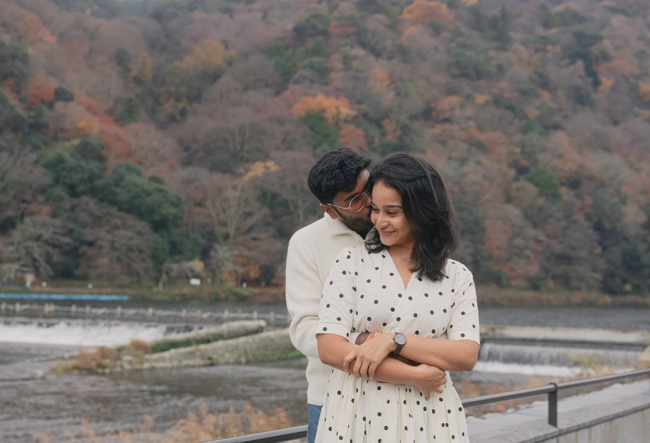 A couple standing near a river with fall foliage in the background. The man is kissing the woman's forehead, both smiling. She wears a white polka dot dress and a wristwatch, he wears glasses and a cream sweater.