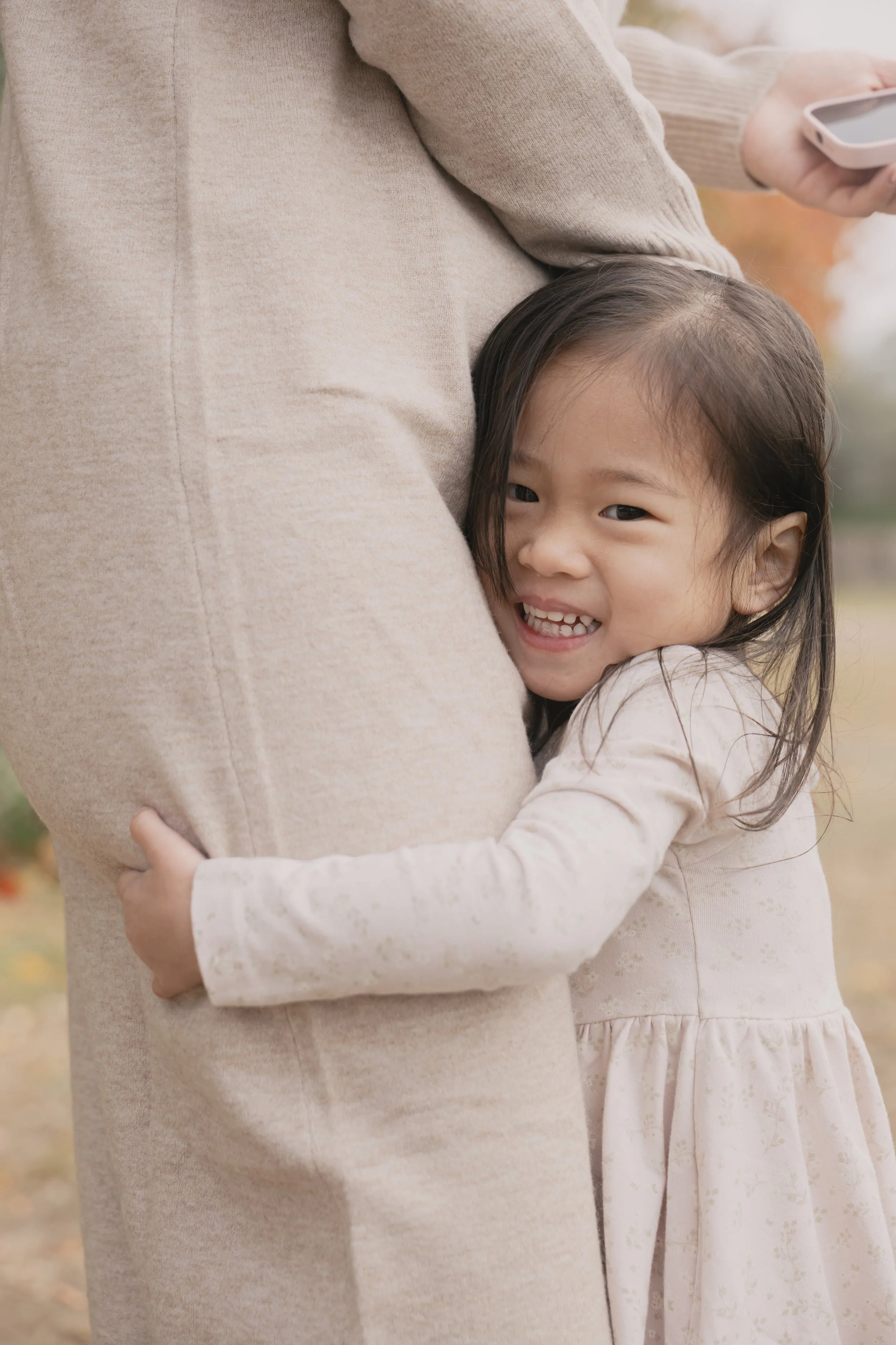 A young girl with dark hair hugging an adult, partially visible, during daytime in an outdoor setting with autumn foliage.