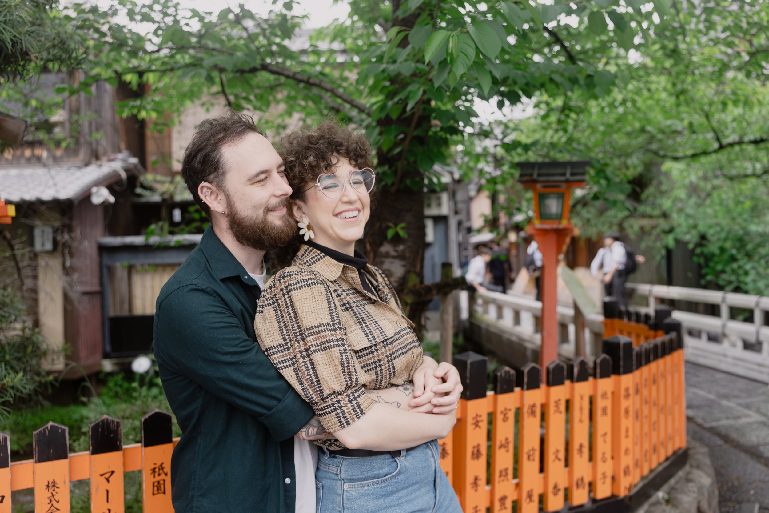 A smiling couple, a man with a beard and earrings and a woman with curly hair and glasses, stands outdoors under a green tree with leaves. They are standing in front of a black and orange fence with Japanese characters on it, near a traditional build