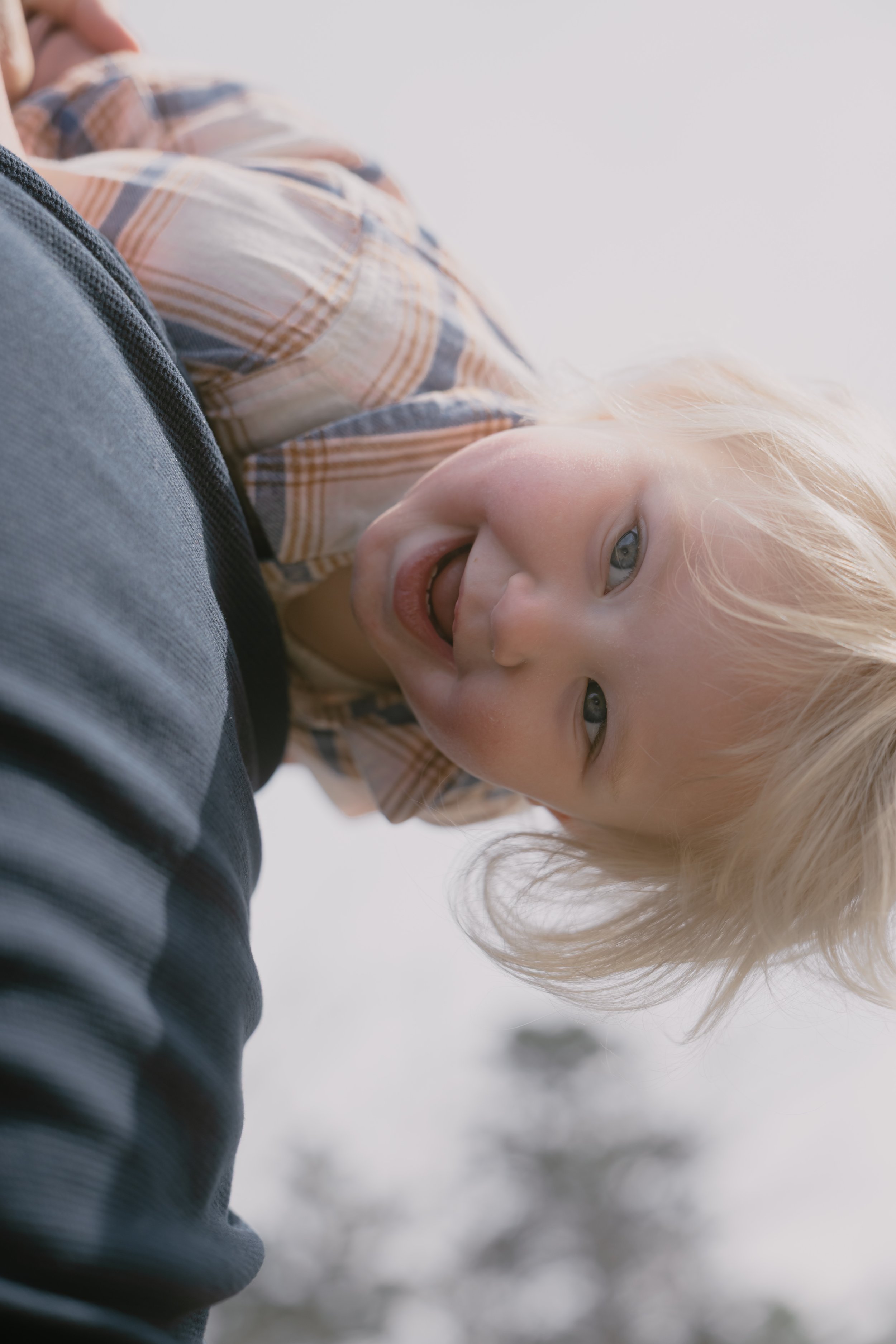 A young child with blonde hair and blue eyes smiling and leaning against an adult's leg, wearing a plaid shirt, outdoors with a blurred sky background.