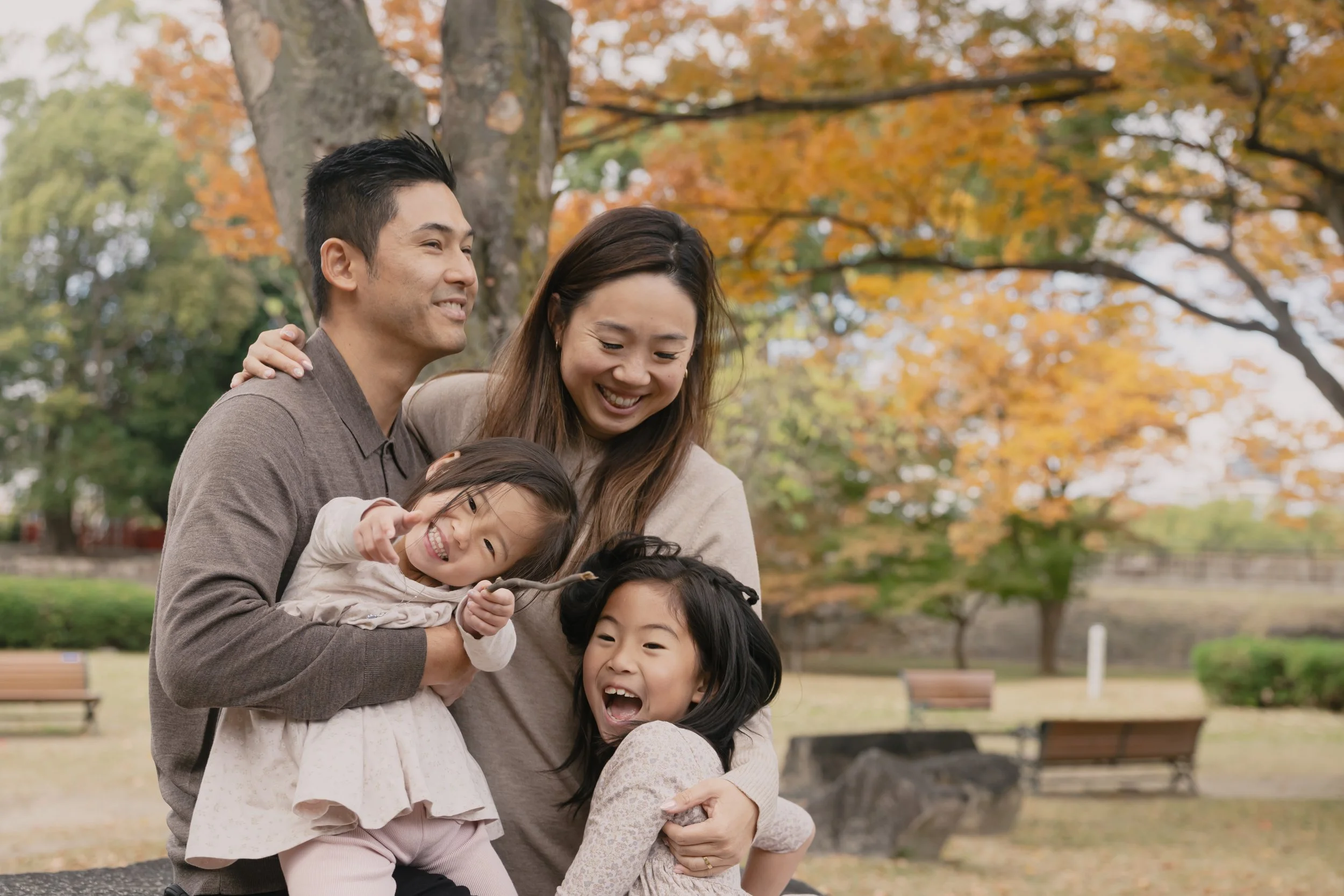 A family with three children and two adults enjoying a fall day in a park. The family is smiling and laughing with autumn trees and benches in the background.