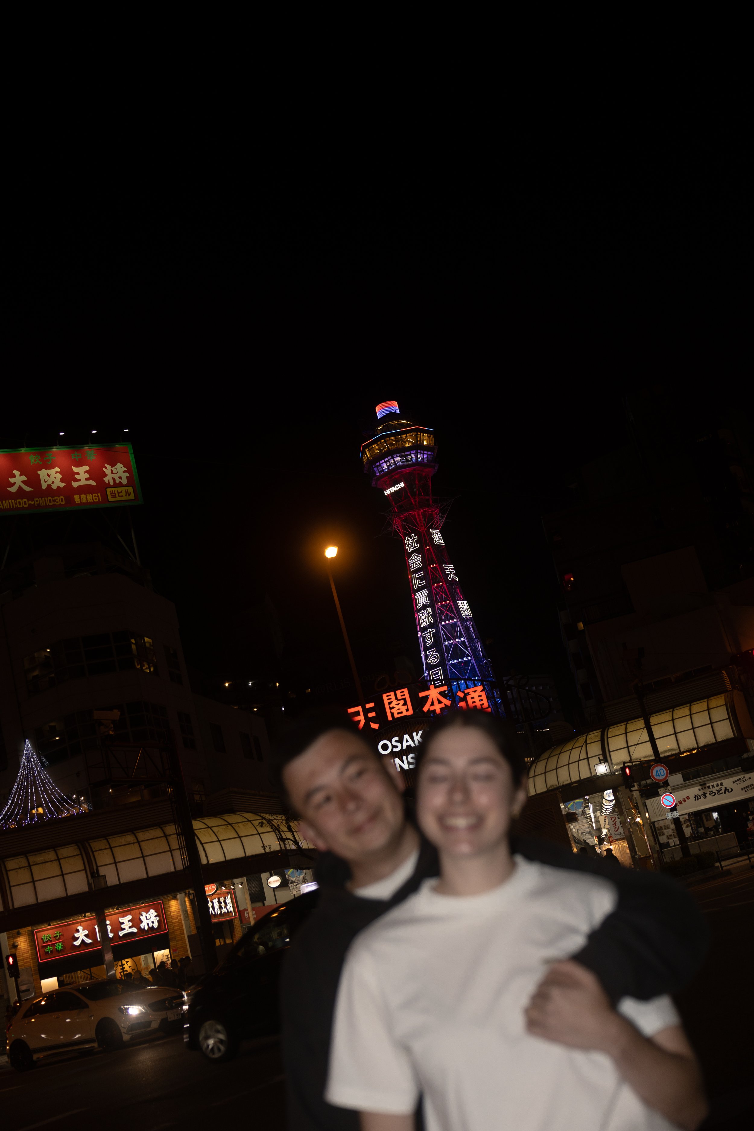 Night scene of a busy urban area with illuminated signage in Japanese characters, a tall lit tower, and two smiling people in the foreground.