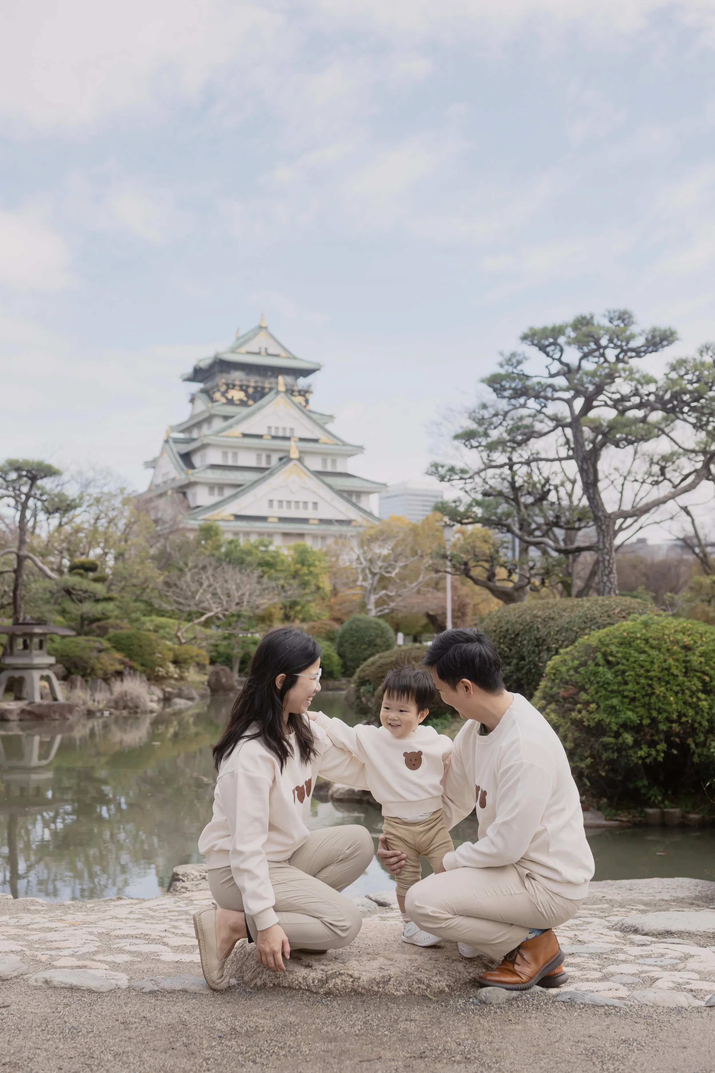 A family of three with a young child outdoors near a pond in a Japanese garden, with a traditional Japanese castle in the background and trees around them.
