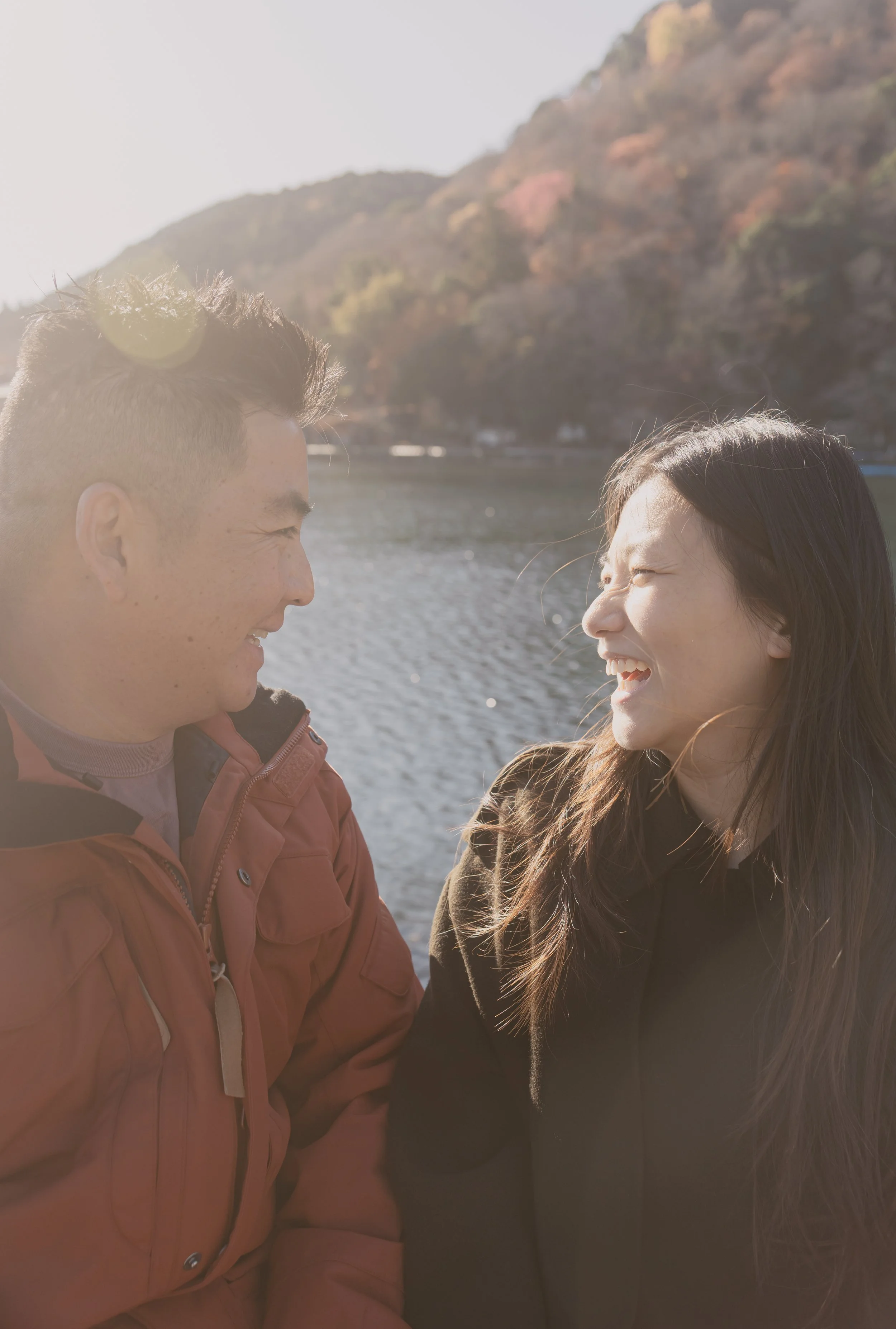 A young man and woman face each other and smile outdoors near a body of water with a hill in the background.