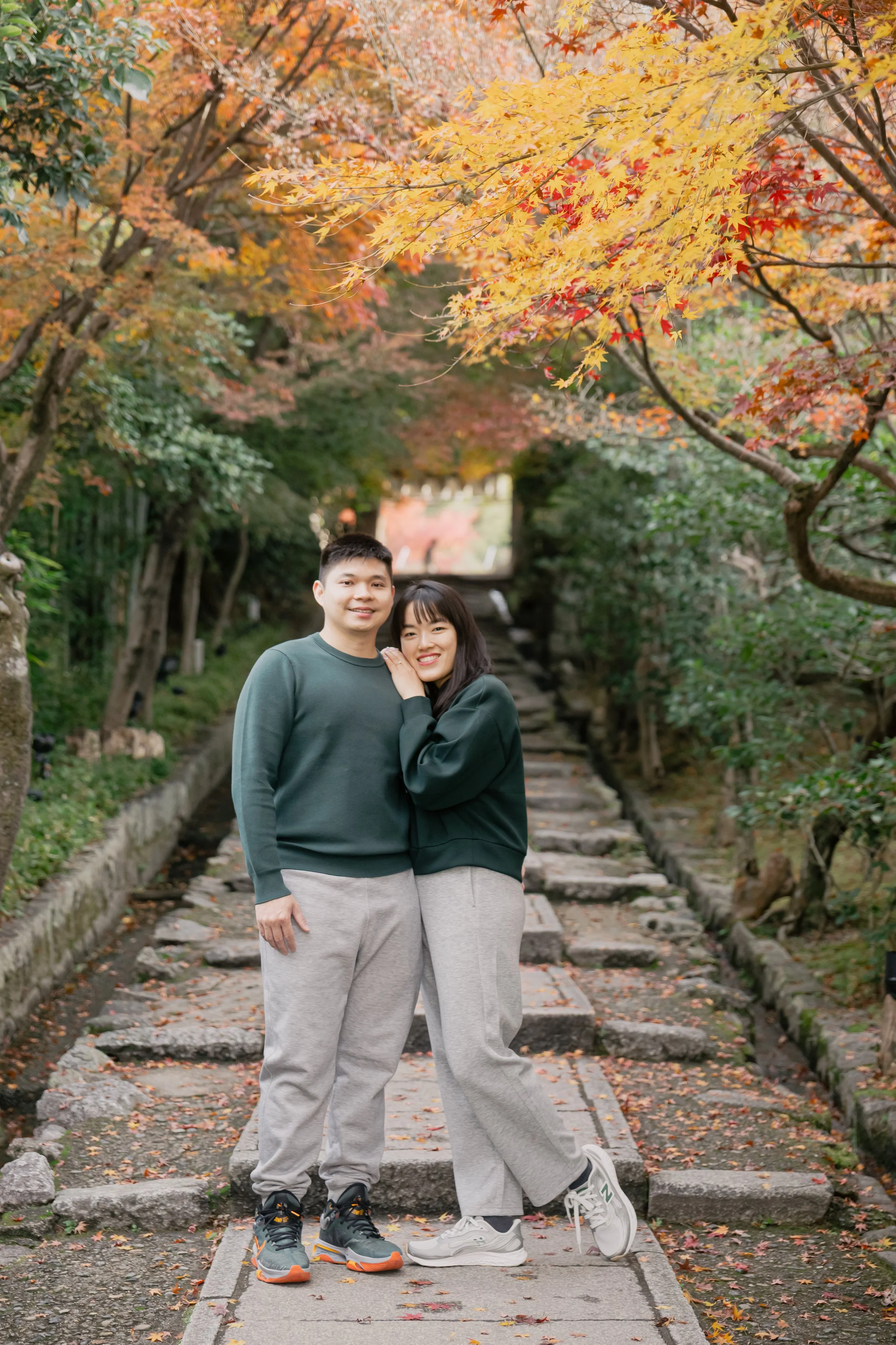 A young couple standing together outdoors on stone steps surrounded by autumn trees with colorful leaves.