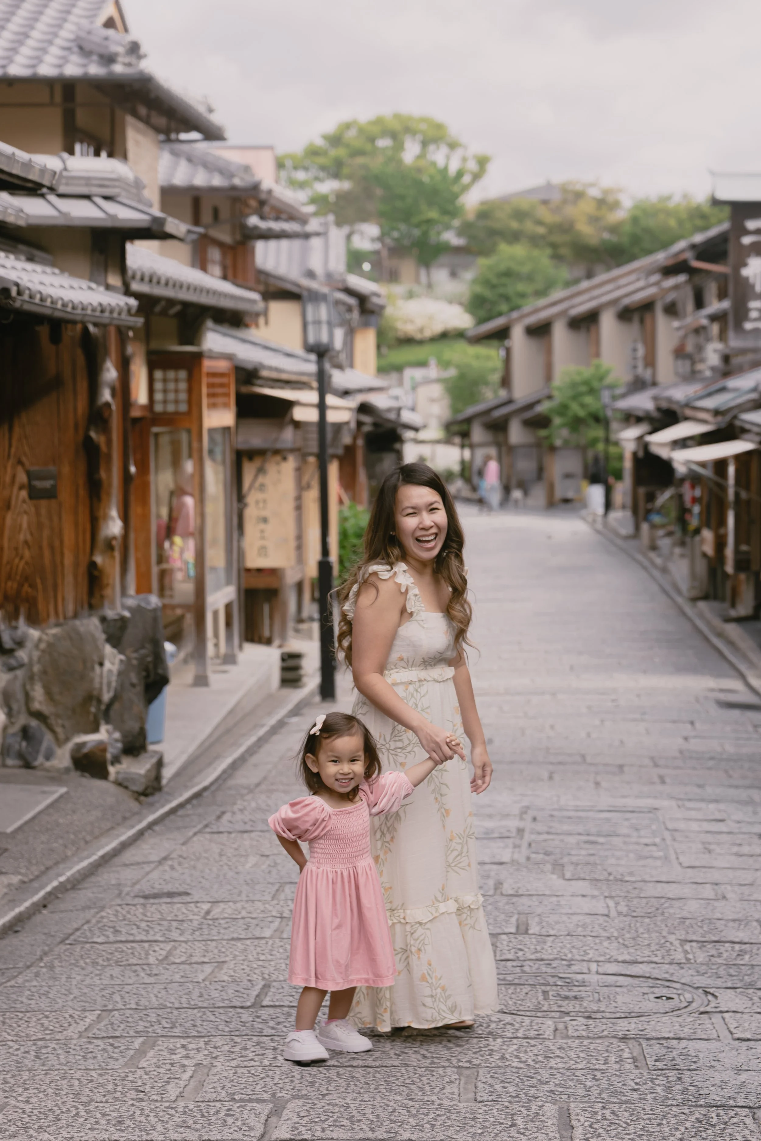 A young woman and a little girl holding hands and smiling on a cobblestone street in a traditional Japanese town with wooden shop fronts and tiled roofs.
