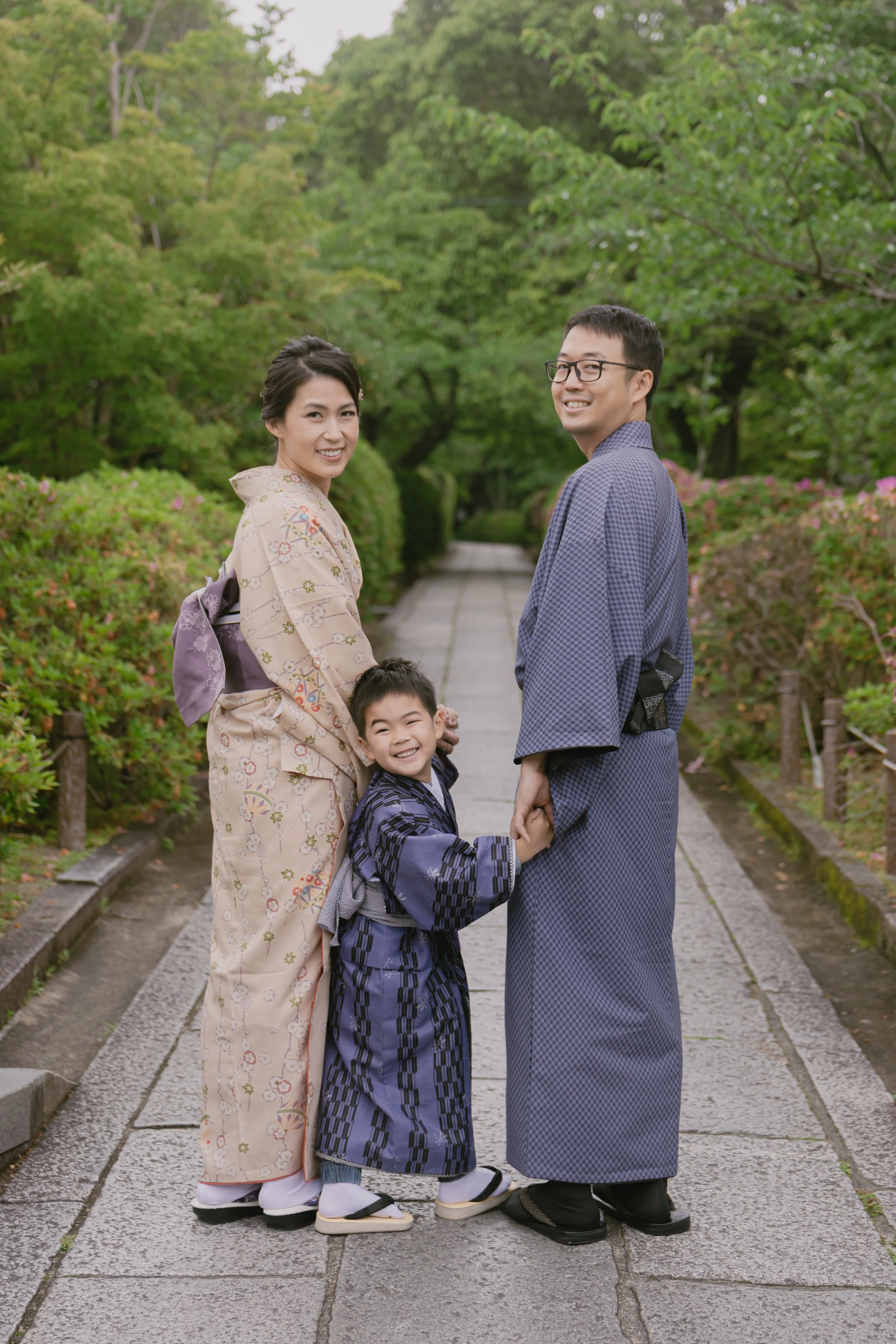 A family of four dressed in traditional Japanese kimonos standing hand in hand and smiling on a garden path surrounded by lush green trees and pink flowers.