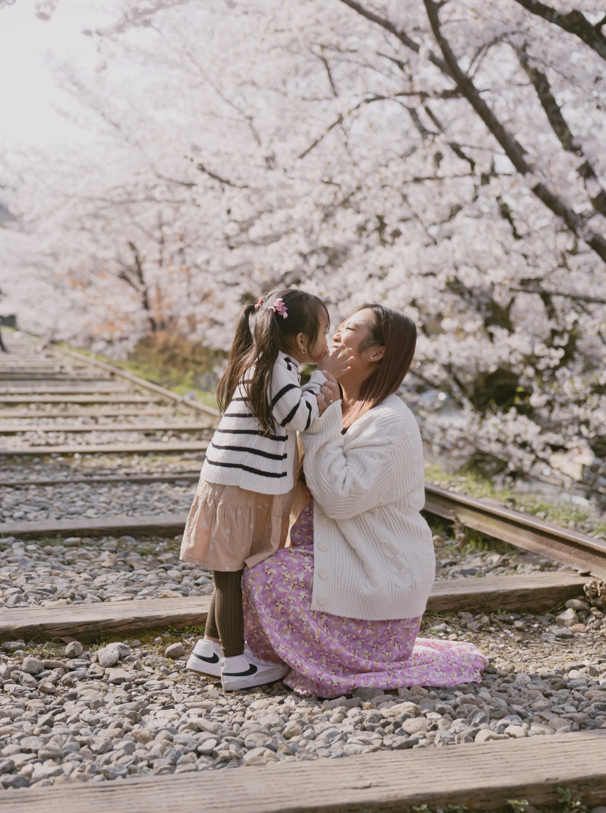 A woman and a young girl sharing a kiss in front of blooming cherry blossom trees near train tracks.