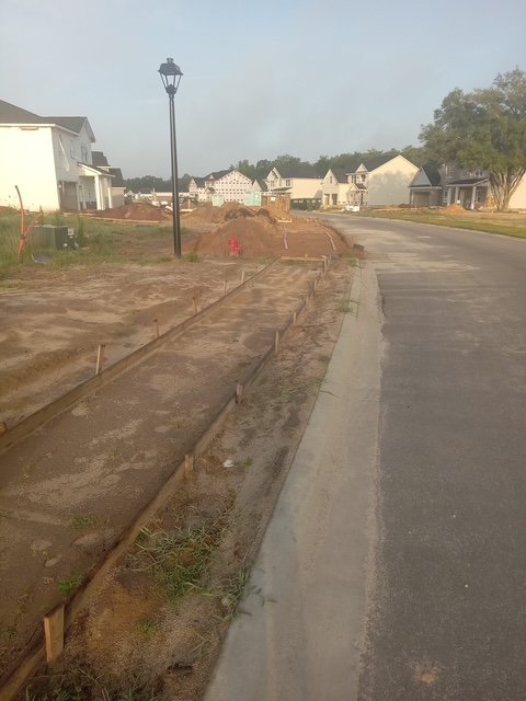 Sidewalk under construction in a residential neighborhood with new houses. The area has unpaved paths marked for concrete. Some houses in the background are partially built.