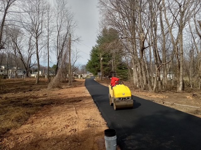 A person operating a small yellow road roller compacts freshly laid asphalt on a rural path