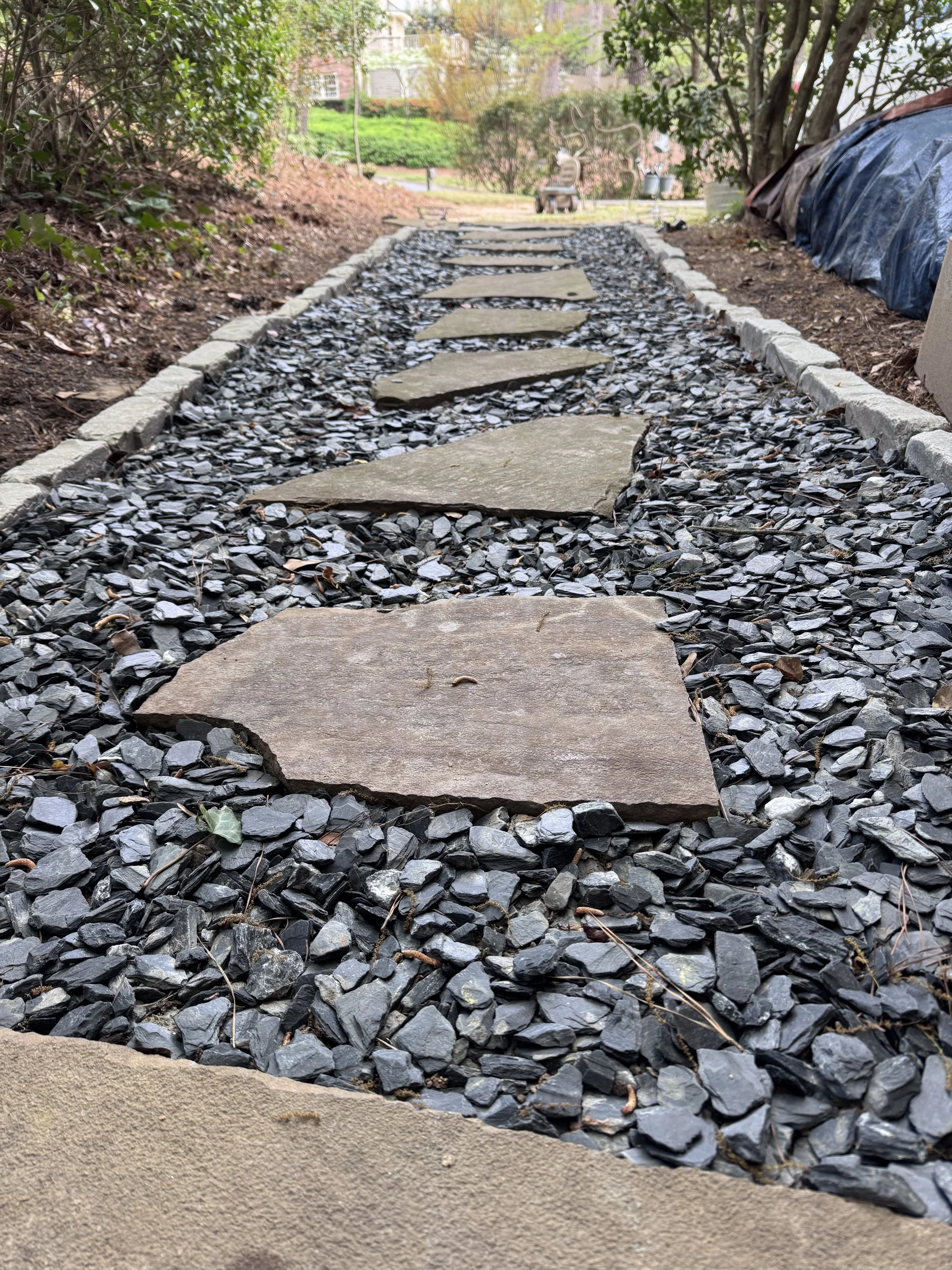 Close-up view of a gravel pathway with large, flat stone stepping stones, surrounded by trees and greenery.