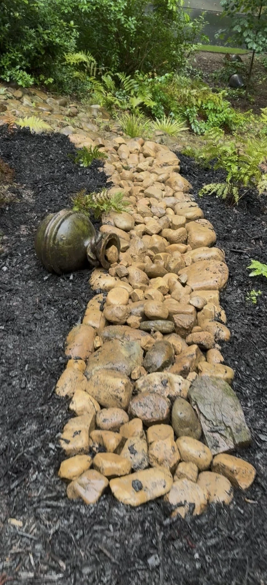 A garden pathway made of tan and brown rocks with black soil on either side, and a fallen decorative ceramic urn on the left side.