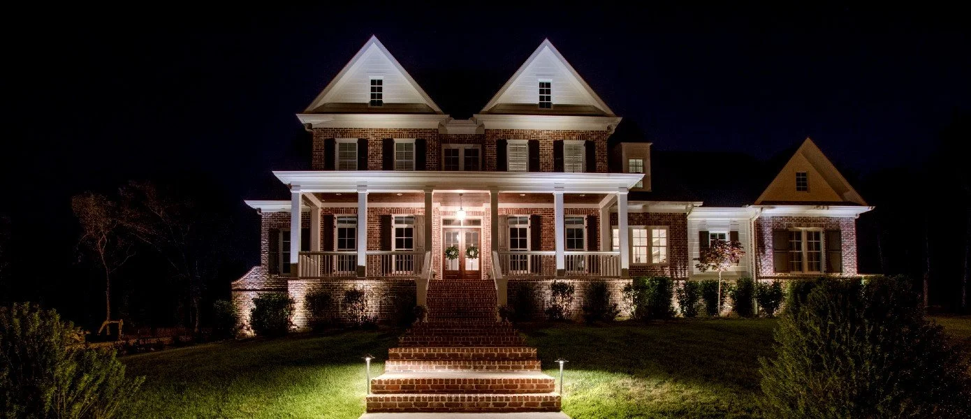 Large brick house at night with illuminated porch, front steps, and surrounding landscaped yard.