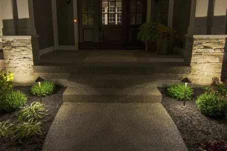Front entrance pathway with stone steps, two small plants, and exterior lighting at night.