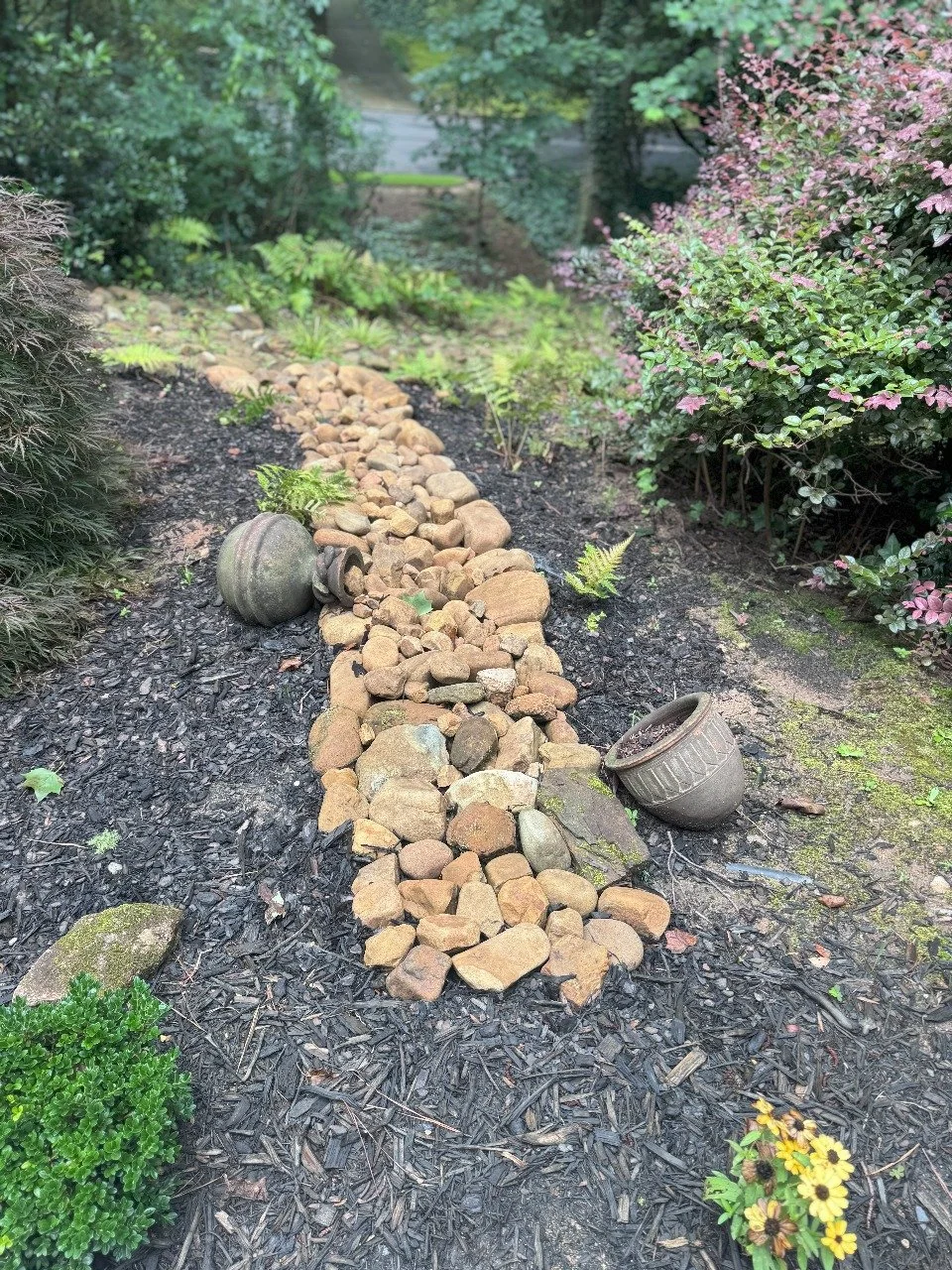 A garden pathway made with small and large rocks, surrounded by various plants and bushes, including a pink flowering shrub, with two upside-down plant pots lying on the ground