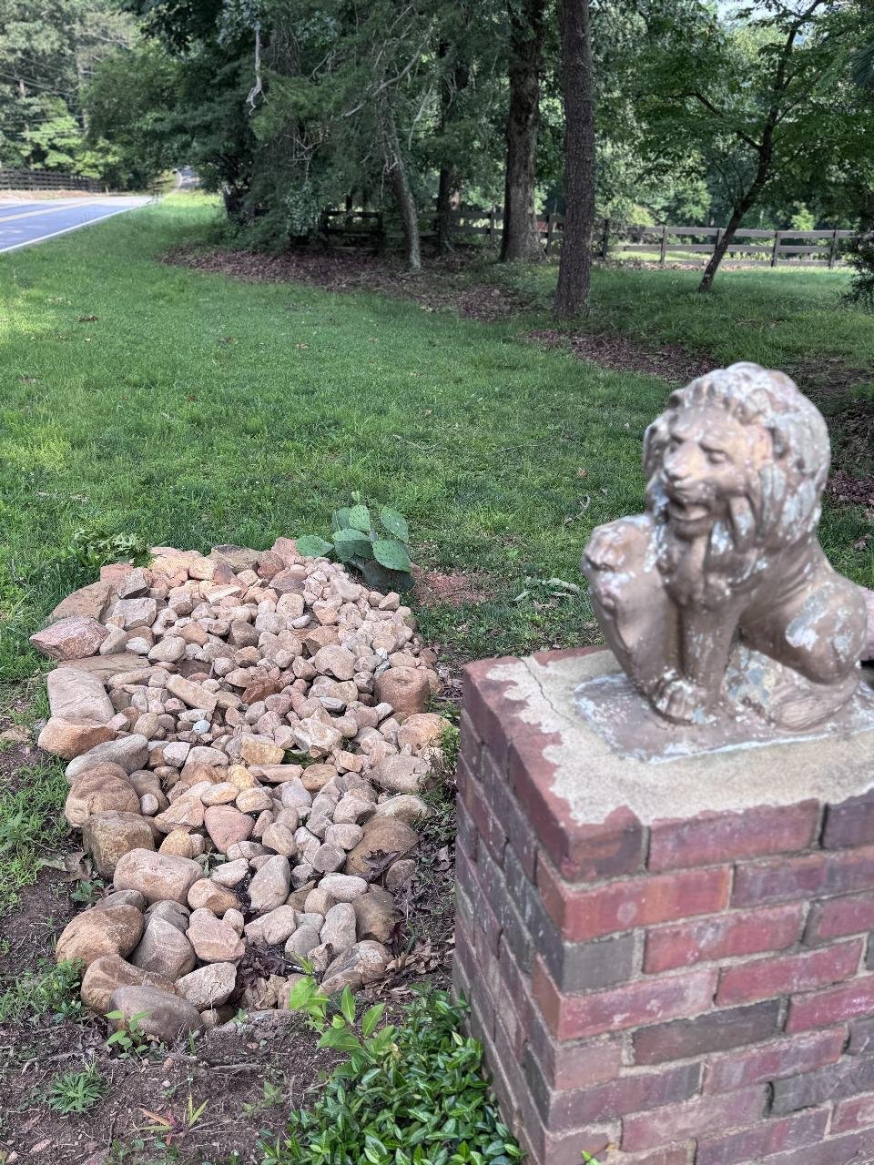 A lion statue on a brick pedestal, a pile of rocks, and some plants in a grassy area next to a road and trees.