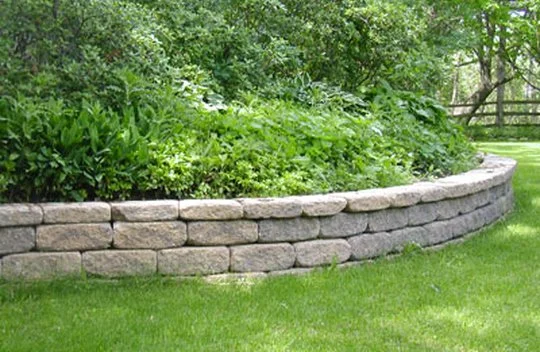 Stone retaining wall with green shrubs and grass in a yard.