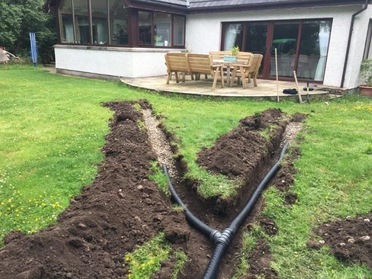 A residential backyard with two trenches dug in the grass, containing black pipes, leading toward the house with an outdoor seating area.