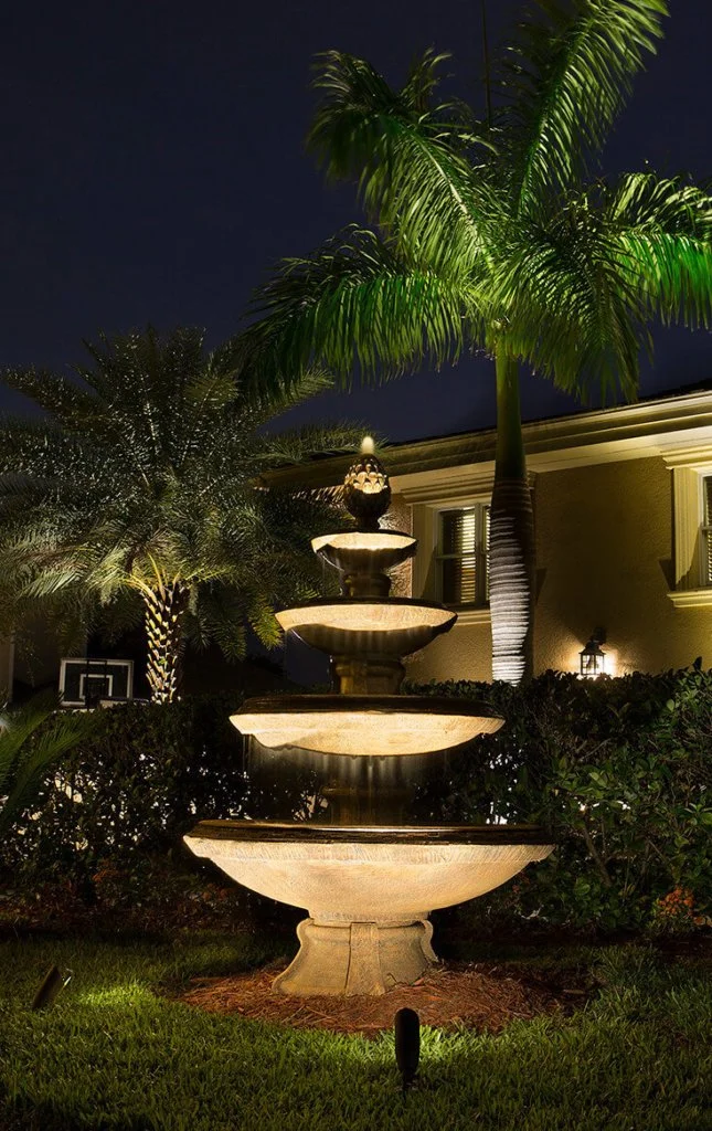 Night view of a lit multi-tiered stone fountain surrounded by landscaped bushes and illuminated palm trees in the yard of a house.