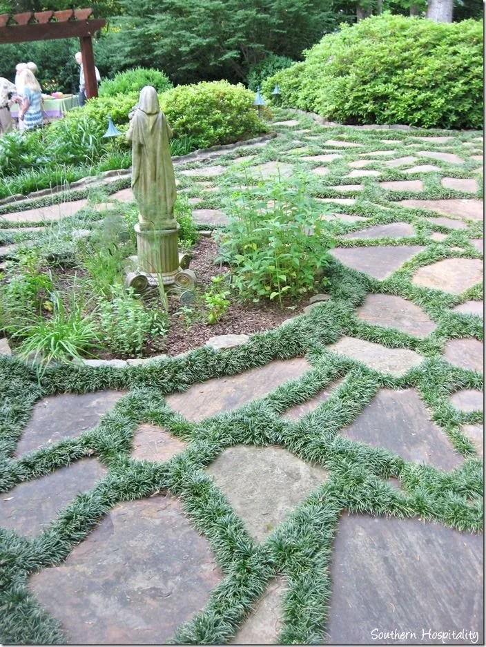Garden pathway with flagstone steps and grass edging, a garden statue, and lush green bushes and trees in the background.