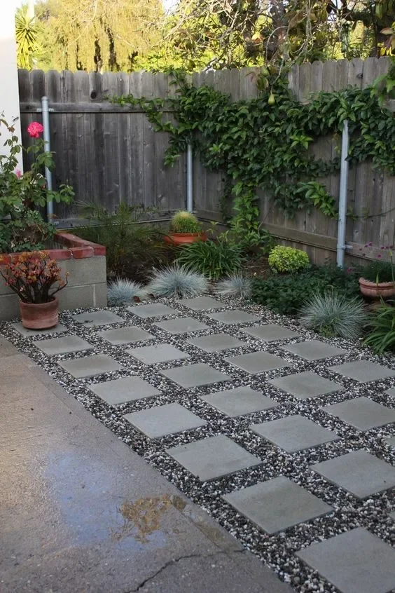A backyard patio with a tiled walkway surrounded by plants and a wooden fence.