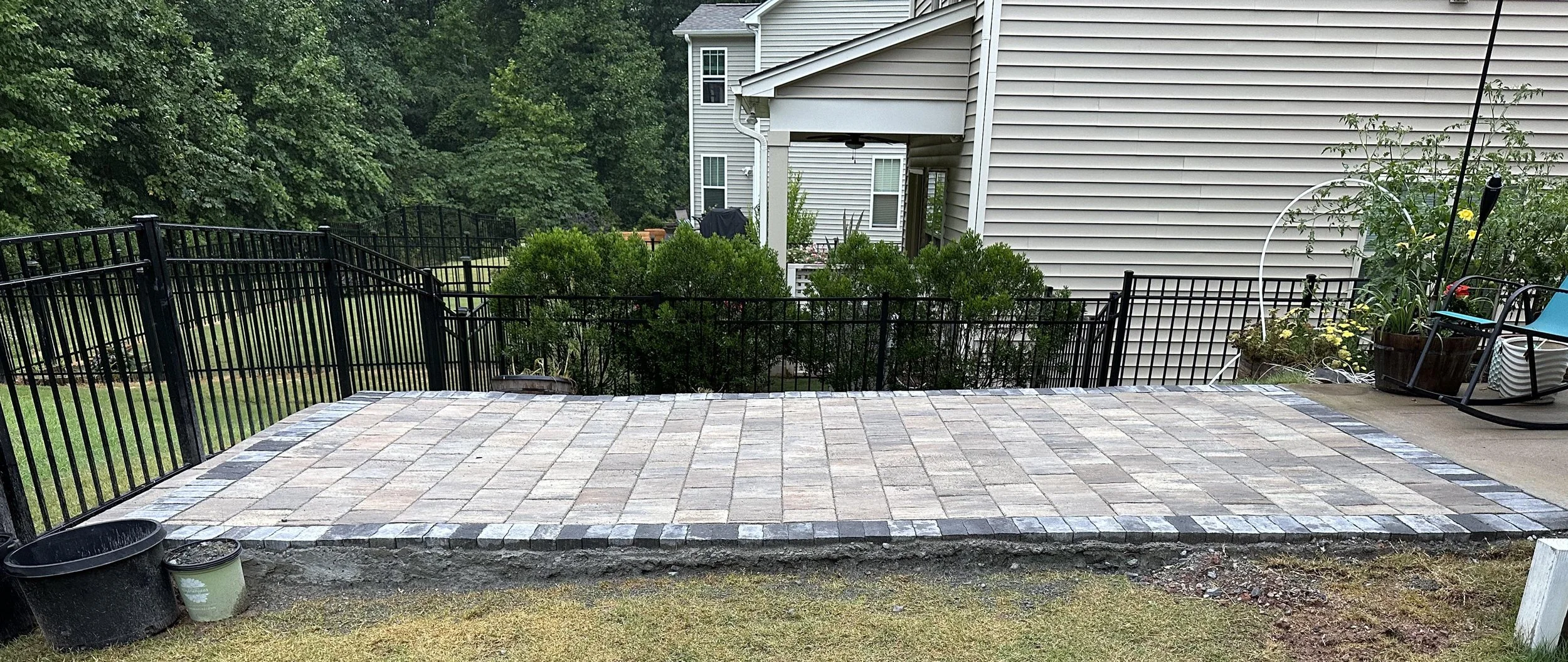 Backyard patio with newly installed stone pavers, surrounded by a black metal fence, with a house in the background and various potted plants and outdoor furniture on the side.