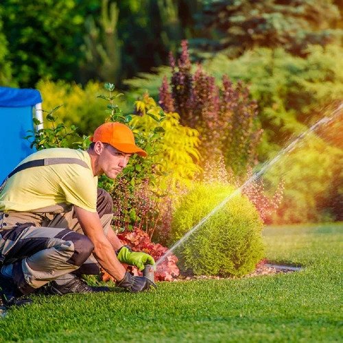 A man wearing an orange cap and yellow shirt watering a garden with a hose in a backyard with colorful plants and trees.