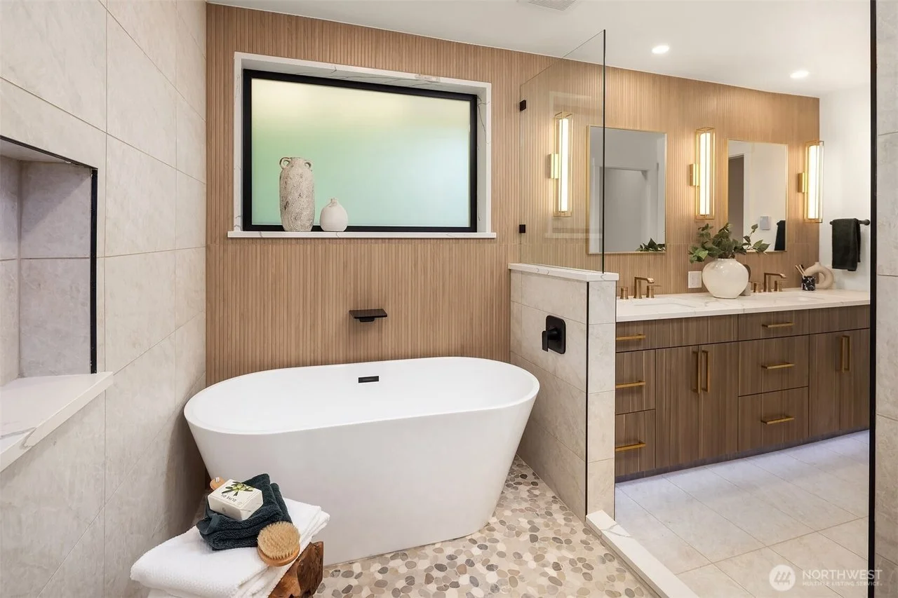 Modern bathroom with a freestanding bathtub, wooden vanity with two sinks, mirror, wall-mounted lights, decorative vases on the window sill, black fixtures, and a pebble-patterned floor near the bathtub.