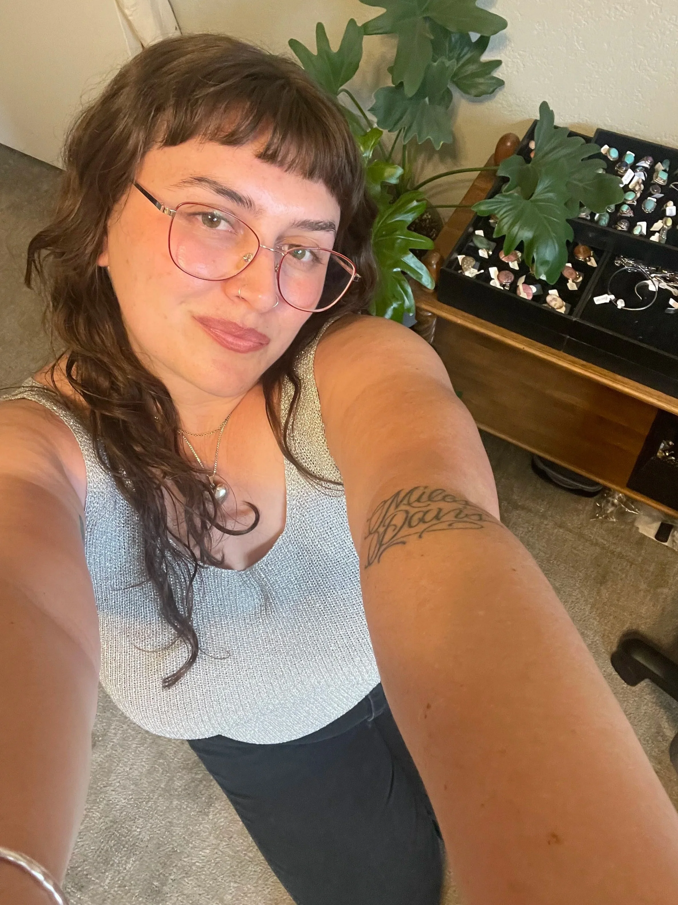 A woman taking a selfie, wearing glasses, a light-colored tank top, and dark pants, sitting next to a wooden table with a large green plant and a jewelry display case filled with various rings and earrings.