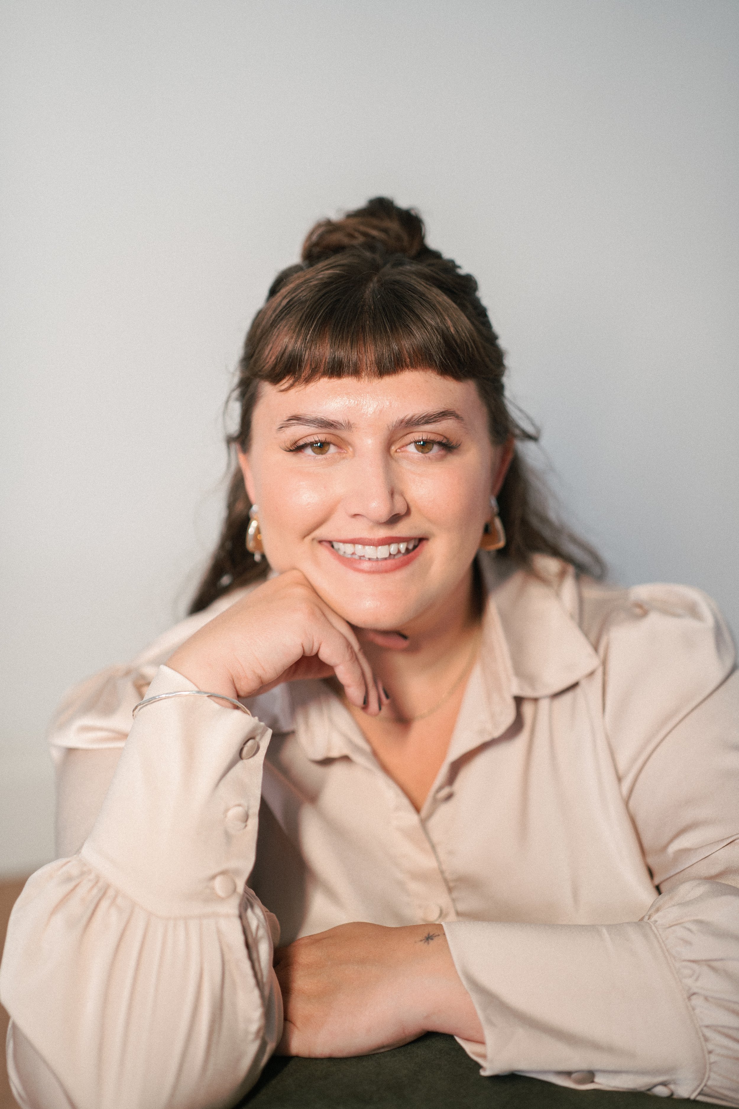 A woman with brown hair styled in a top bun and bangs, wearing gold earrings, smiling with her cheek resting on her hand, wearing a beige blouse with puffed sleeves, sitting at a table with a plain white wall background.