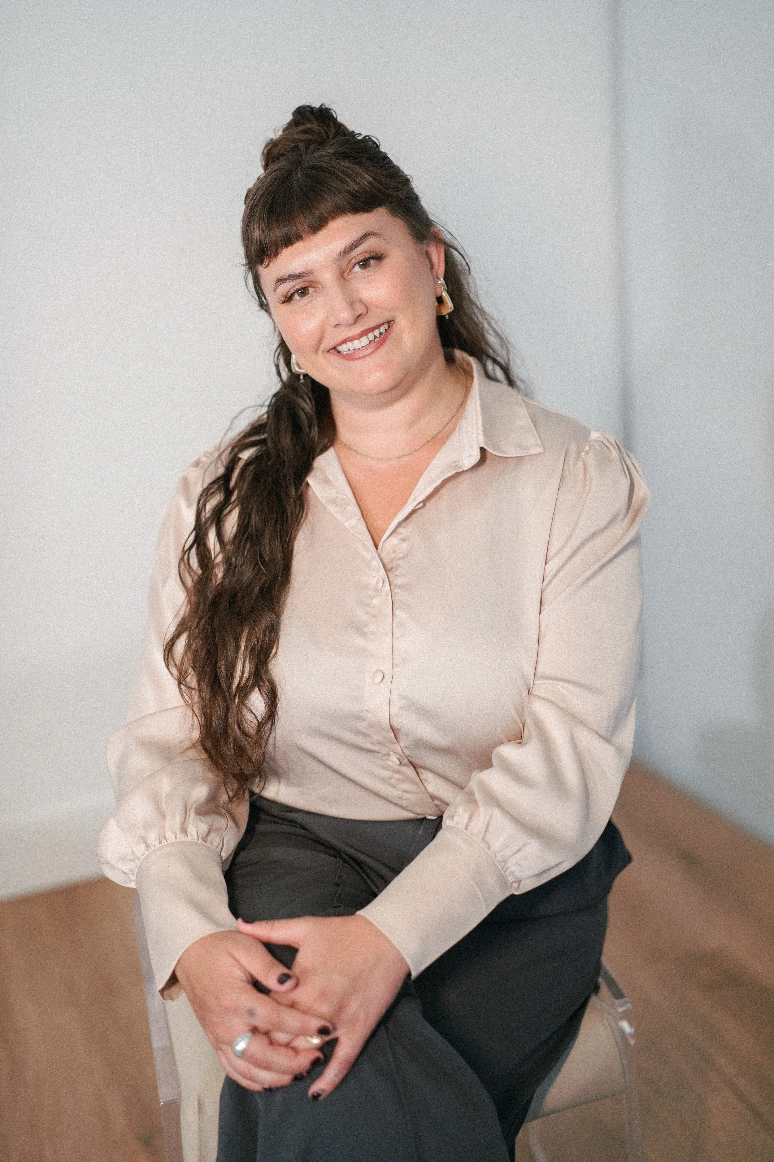 A woman with dark brown hair styled in a half-up, half-down with blunt bangs, sitting on a chair indoors, smiling at the camera, wearing a beige silk blouse and black pants.