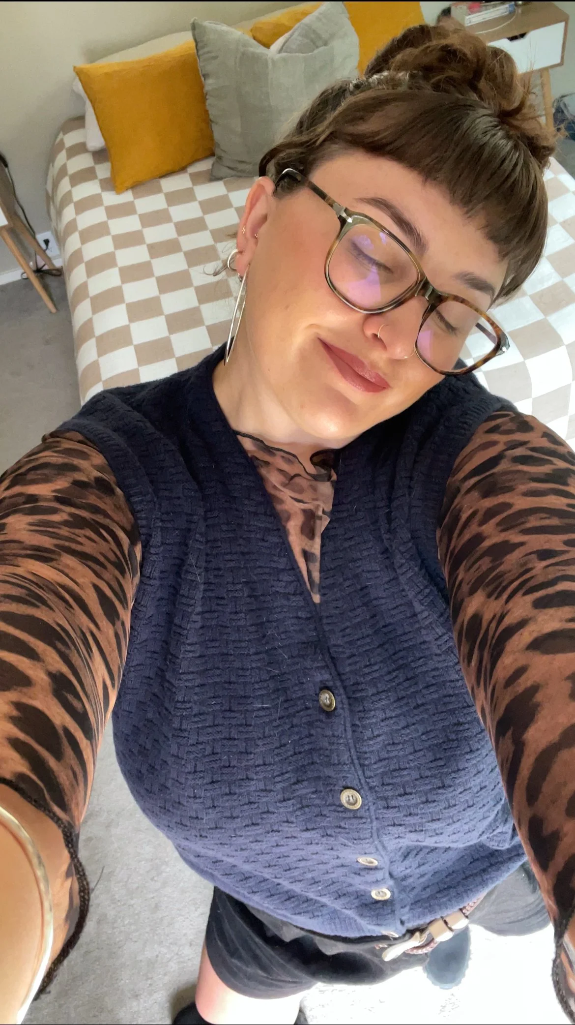 A person taking a selfie in a bedroom, with a checkered bedspread, gray and yellow pillows, and a bedside table visible in the background.