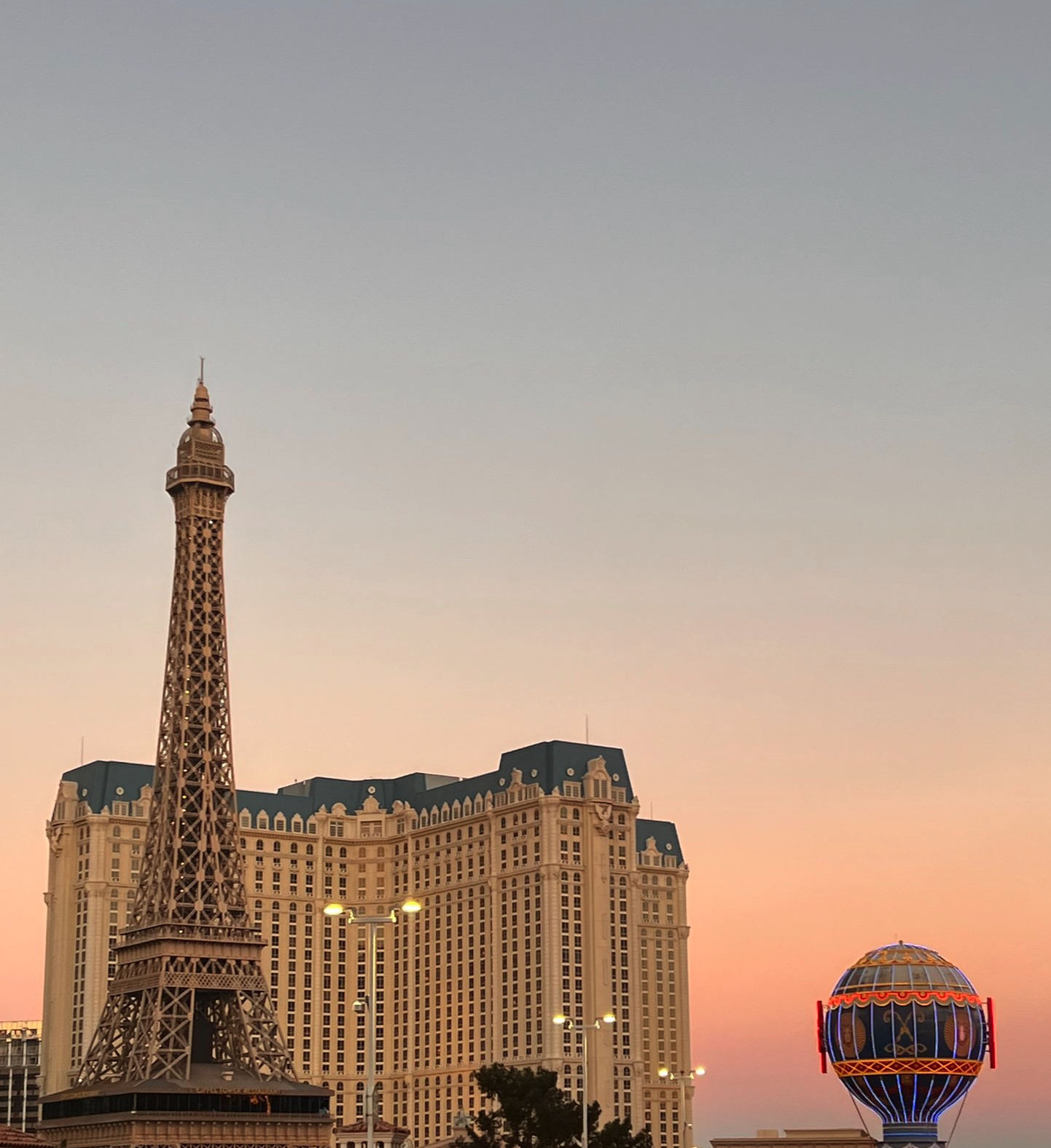 In the foreground, there is a miniature Eiffel Tower replica. Behind it is the Caesars Palace hotel and casino in Las Vegas, with its large beige building and blue-tiled roof. The sky is at sunset, with warm orange and pink hues. To the right, there is a colorful, illuminated hot air balloon-shaped structure.
