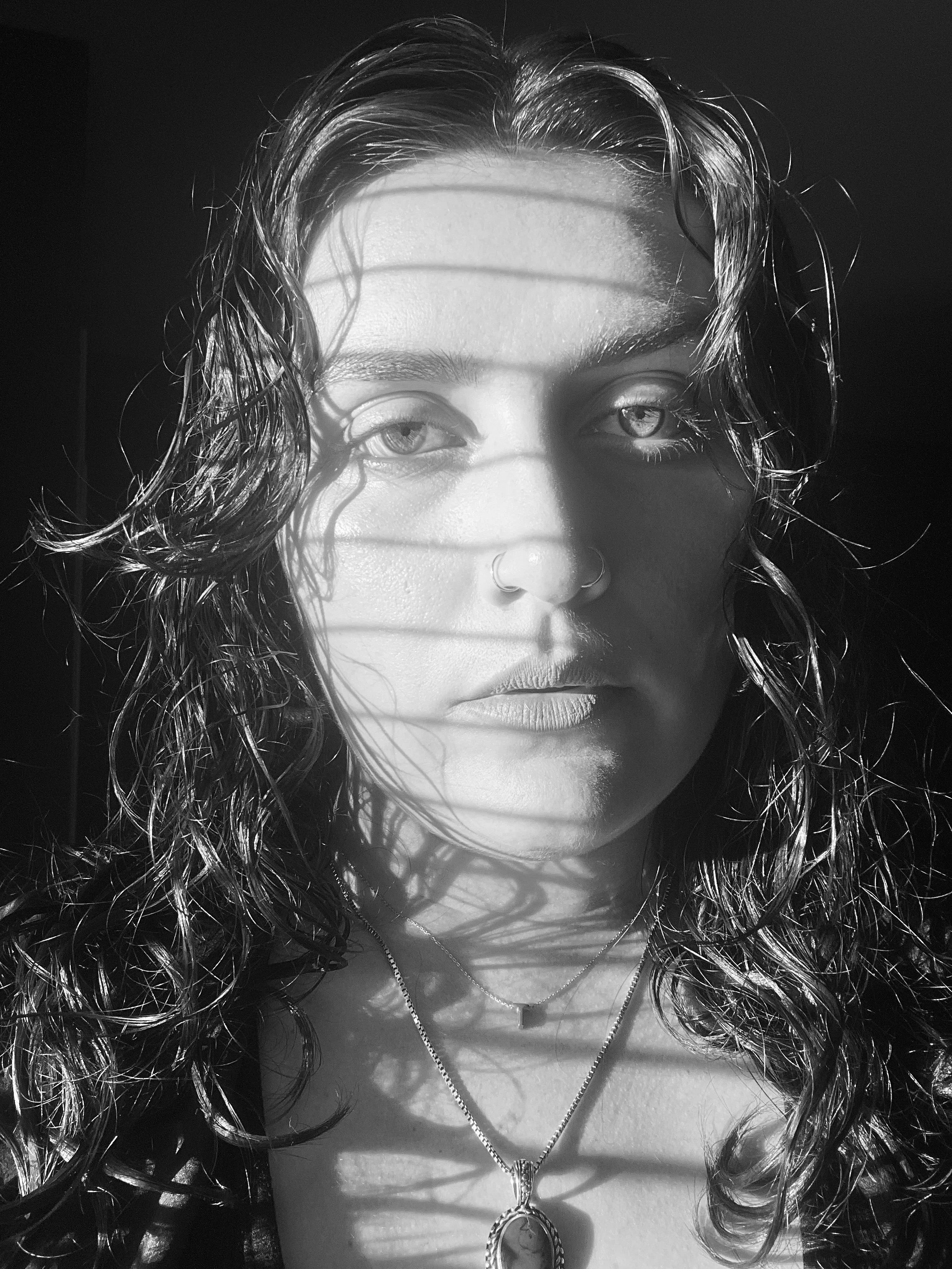 Close-up black and white portrait of a woman with wet, curly hair and light eyes, wearing necklaces and a nose ring.