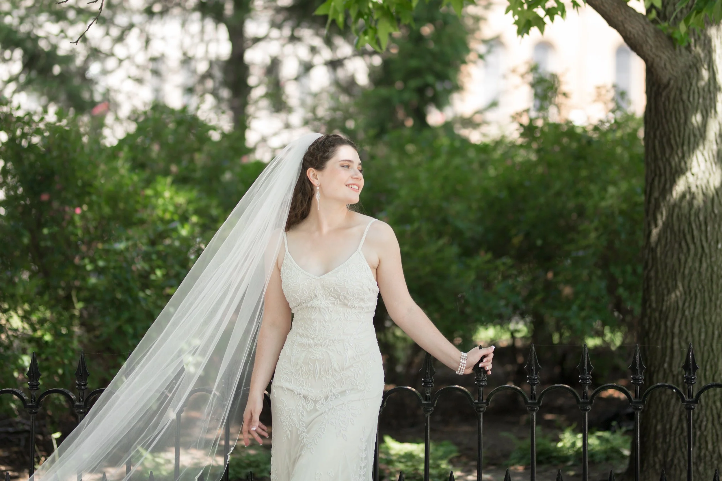 A bride in a white wedding dress with lace details and thin straps, wearing a long veil and jewelry, standing outdoors near a black wrought iron fence and green trees, smiling and looking to her left.