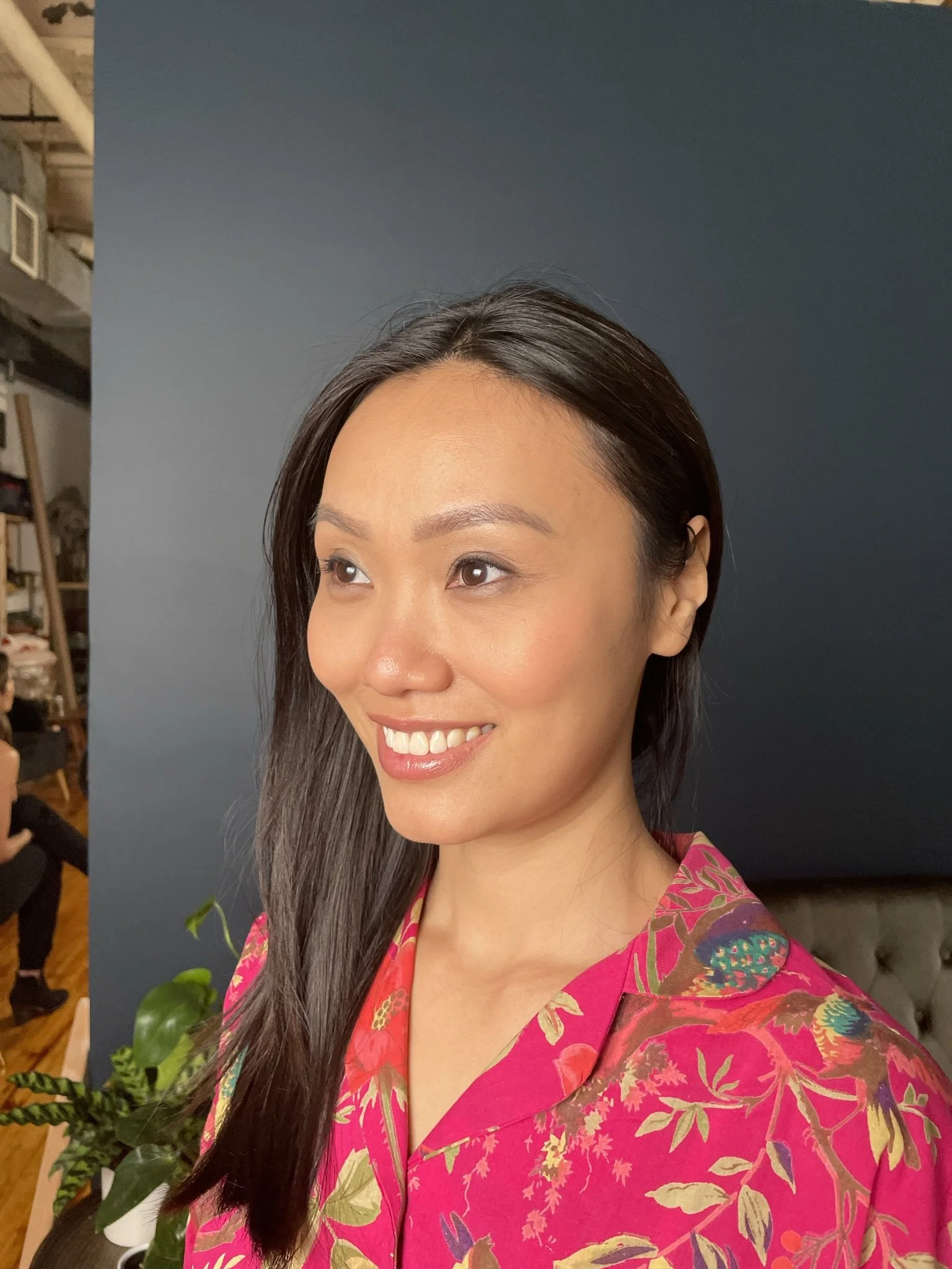 A woman with long dark hair smiling, wearing a pink floral patterned shirt, standing in front of a dark background, with some plants and a person in the background.