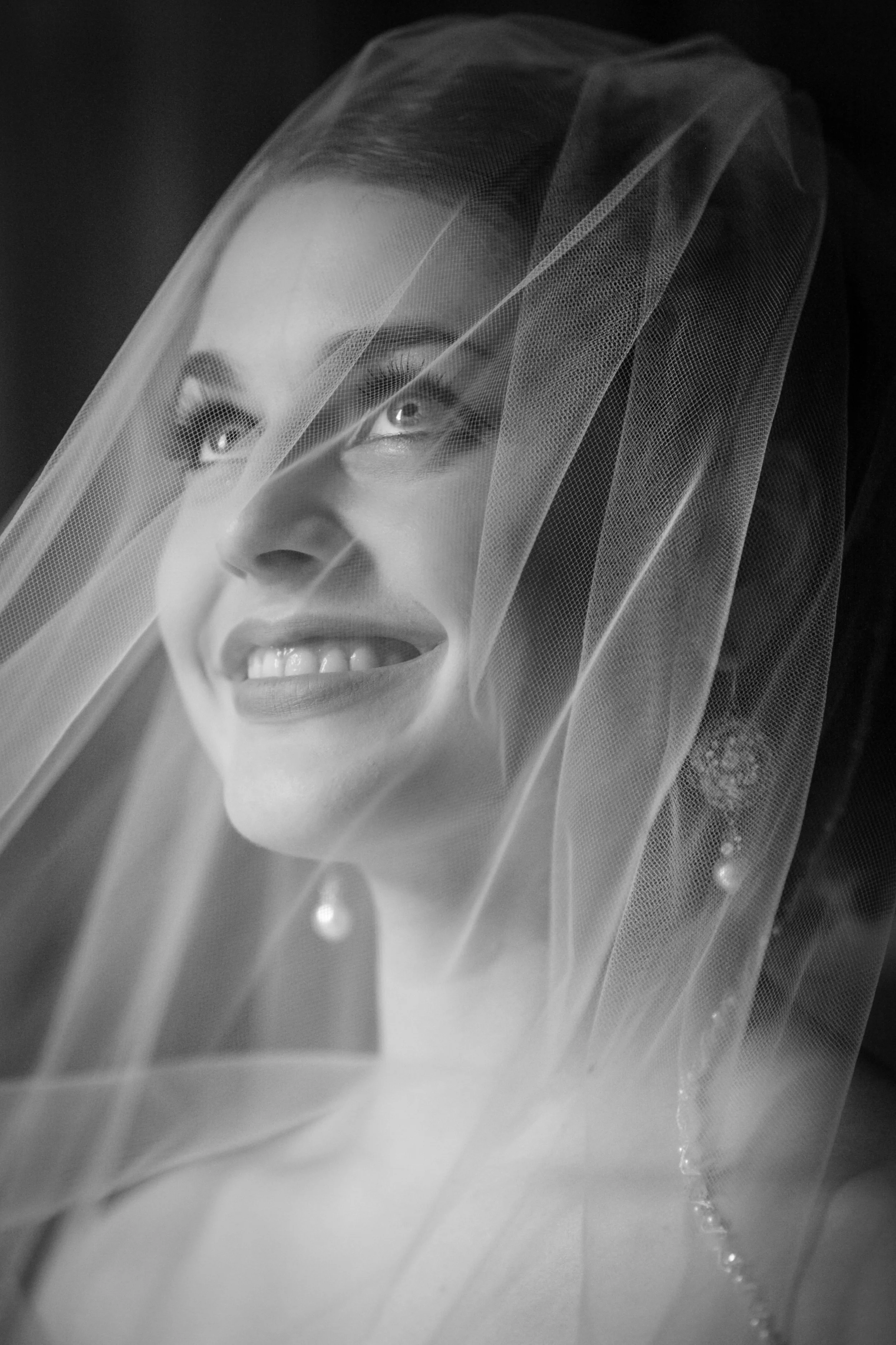 A black-and-white portrait of a woman wearing a bridal veil and pearl earrings, smiling and looking slightly upward.