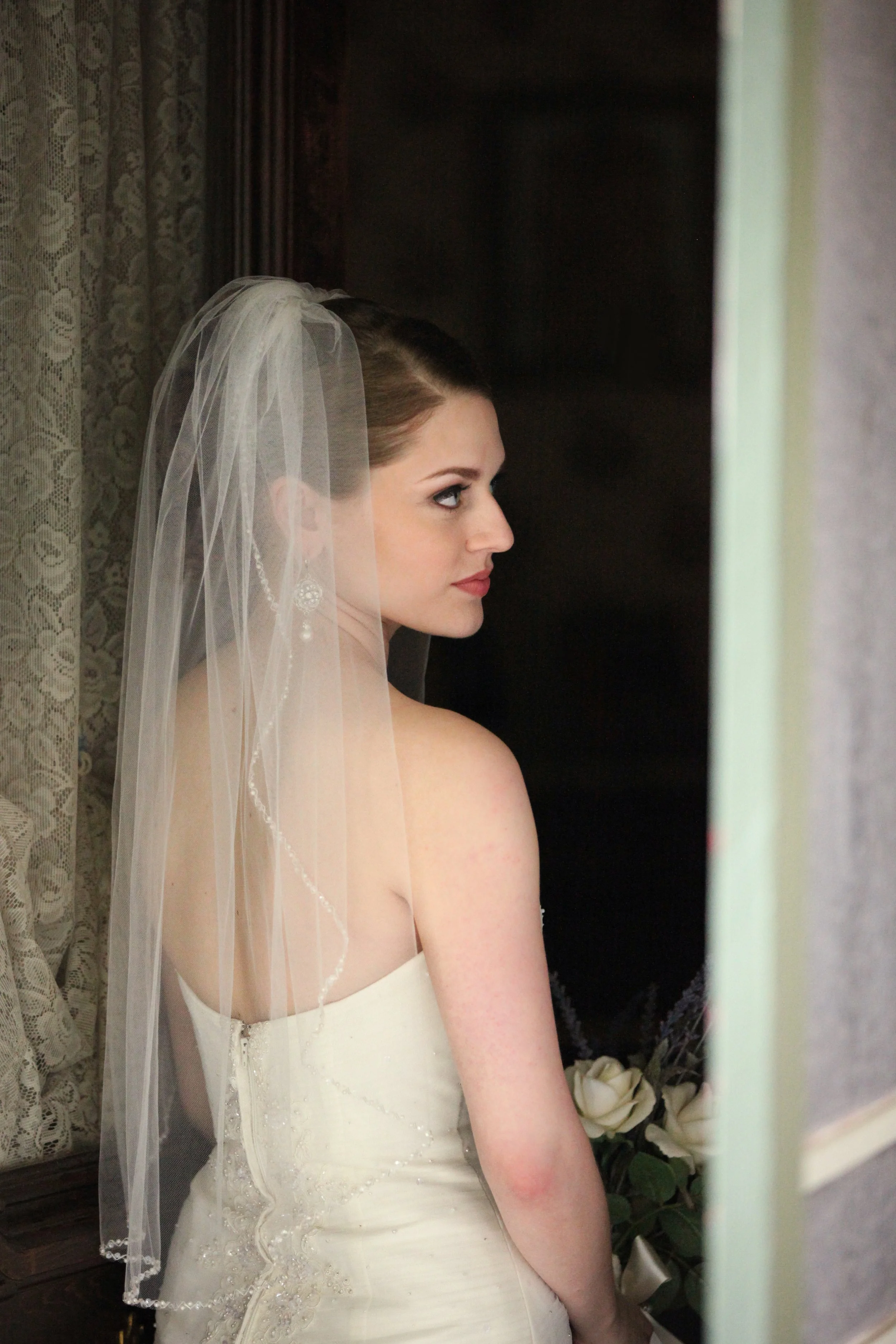 A bride with a sheer veil, wearing pearl earrings and a strapless white wedding dress, stands near a window with lace curtains, holding a bouquet of white roses.