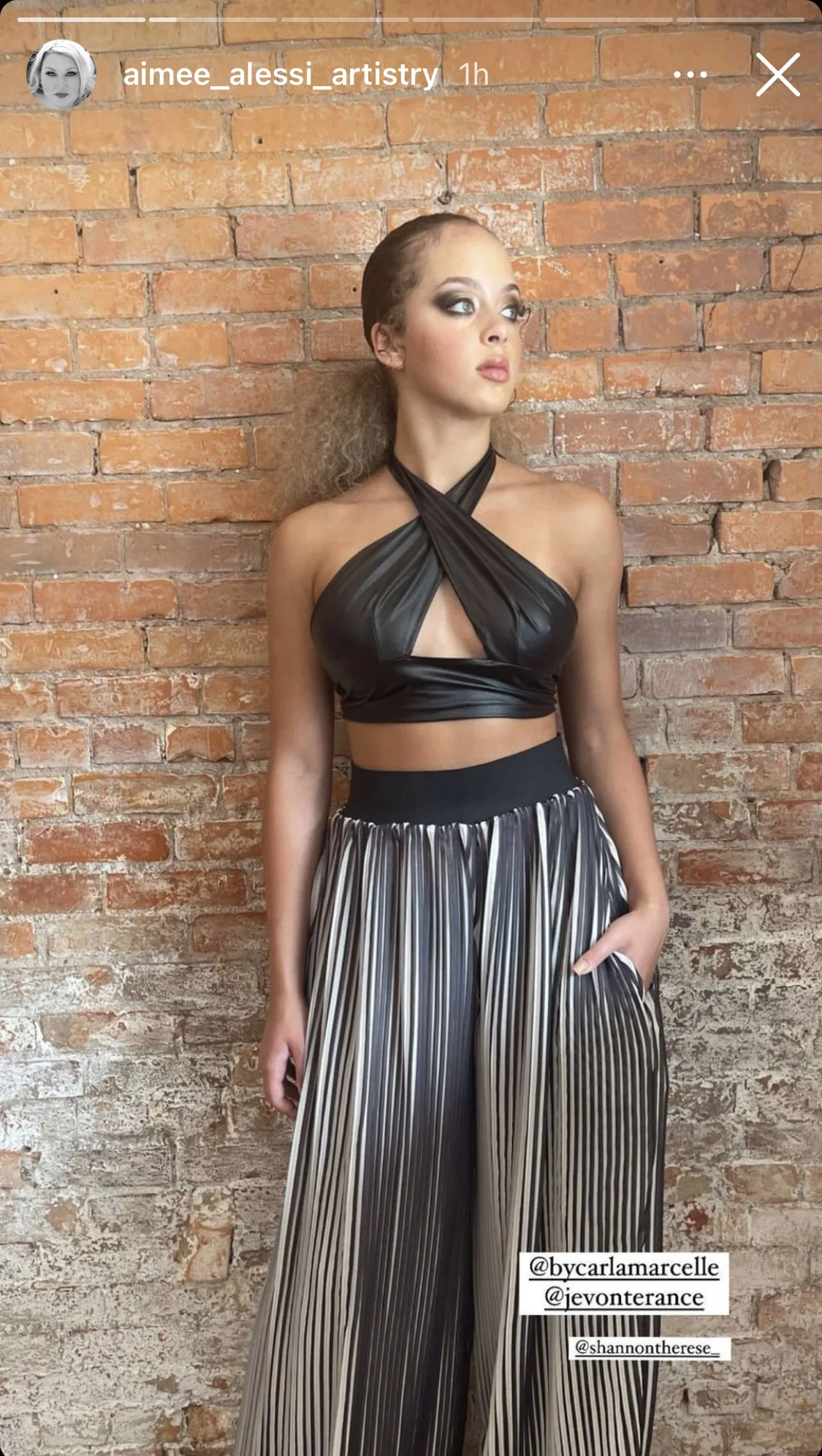 A woman with curly hair styled pulled back, wearing a black halter top with a crisscross design and high-waisted, wide-leg striped pants, standing against a red brick wall.