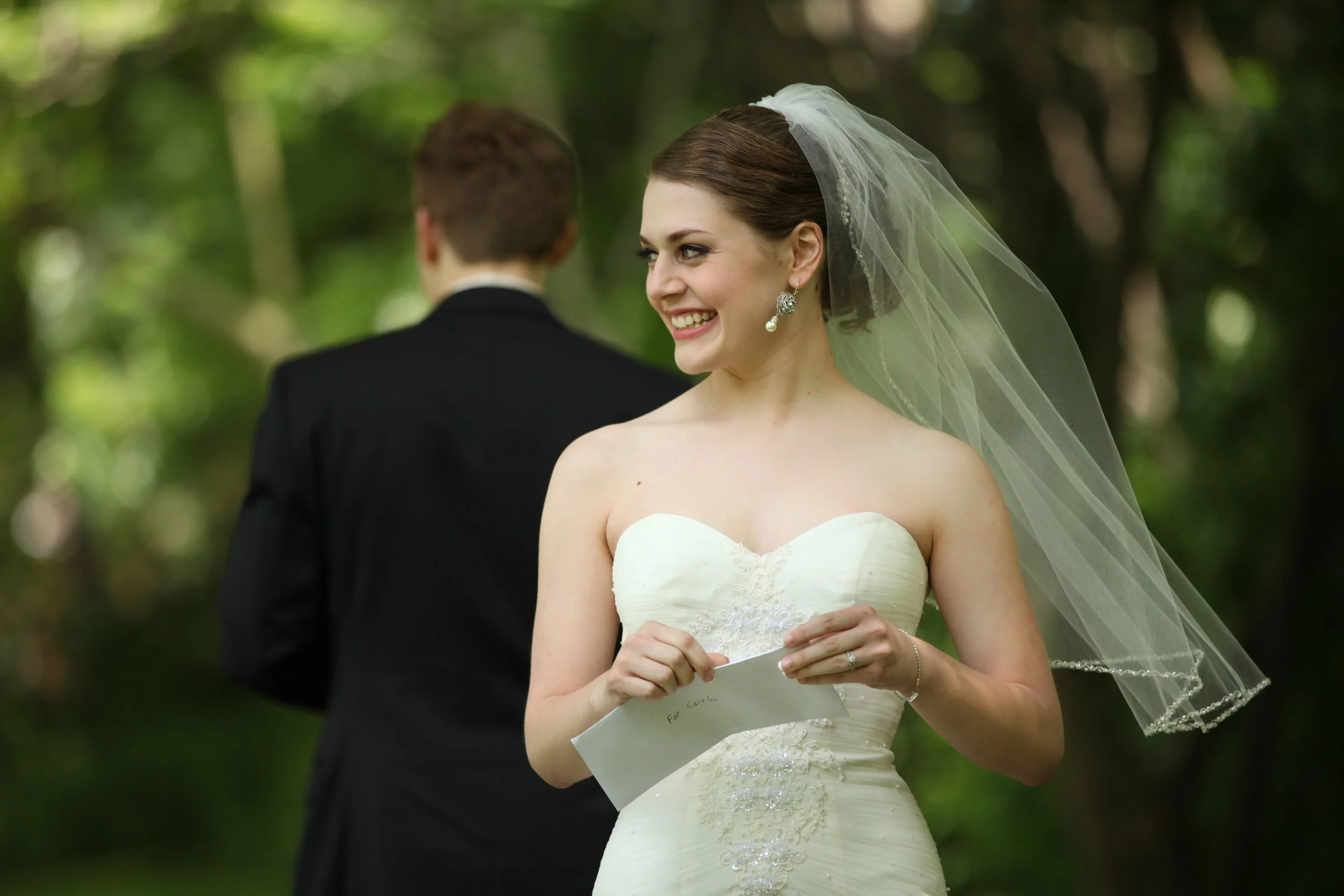 A smiling bride in a strapless white wedding dress with lace detailing, wearing a veil and earrings, holding a small note, with a groom in a black suit standing behind her in a green outdoor setting.